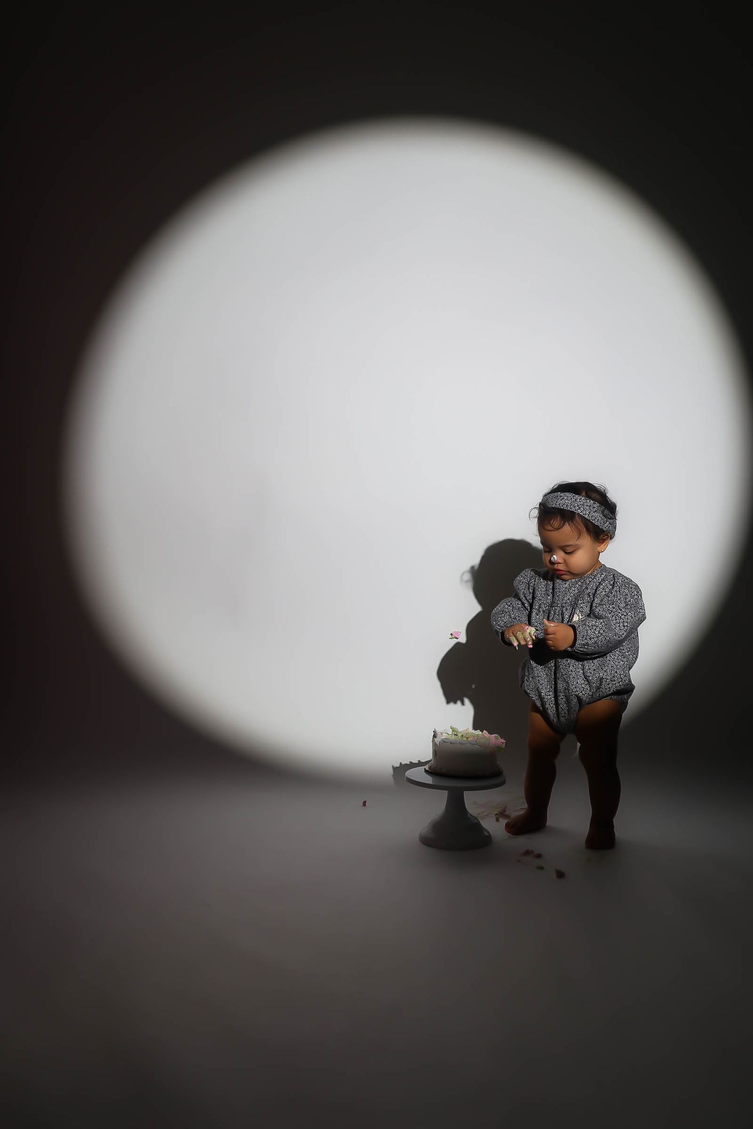 A toddler in a gray outfit stands by a small birthday cake under a dramatic circular spotlight on a dark background.