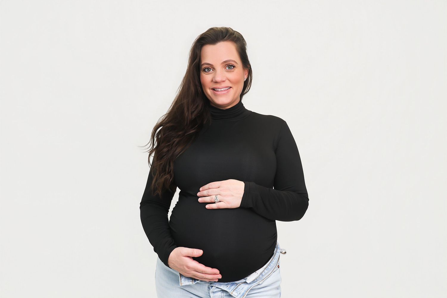 Smiling pregnant woman in black turtleneck and jeans cradling baby bump against white background.