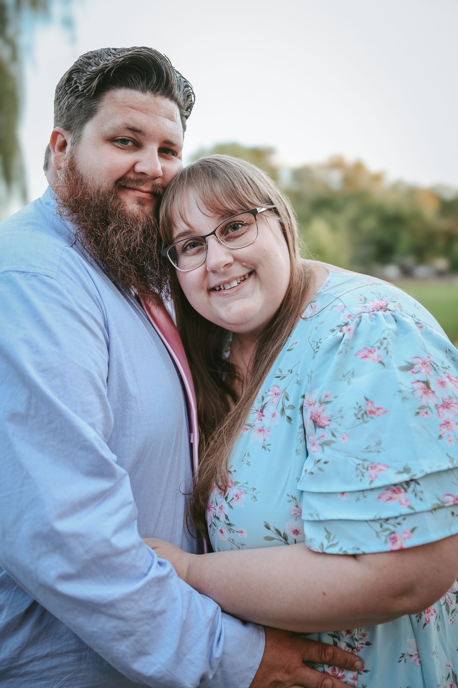 A couple embraces outdoors while wearing a light blue floral dress and white shirt with a full beard.