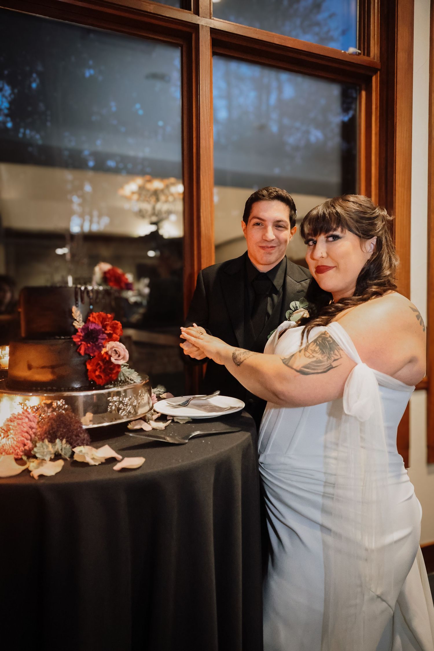 A couple sits at an elegantly decorated wedding reception table with red floral centerpieces and formal place settings.