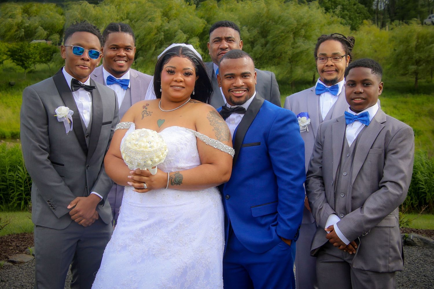 Wedding party in blue and gray suits poses with bride in white dress outdoors in garden setting.