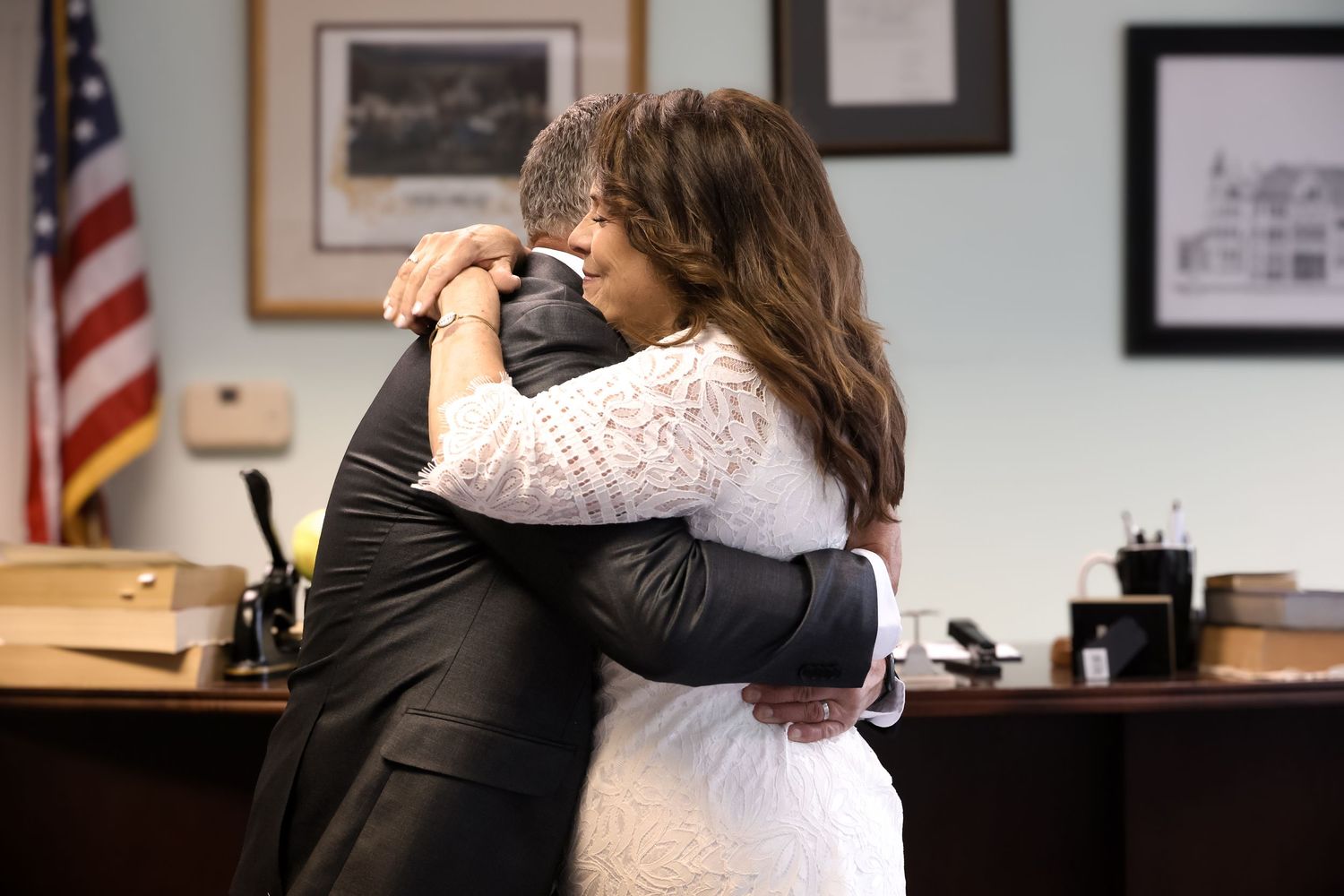 Two people embrace in an emotional hug inside an office setting with American flags and framed photos on the wall.