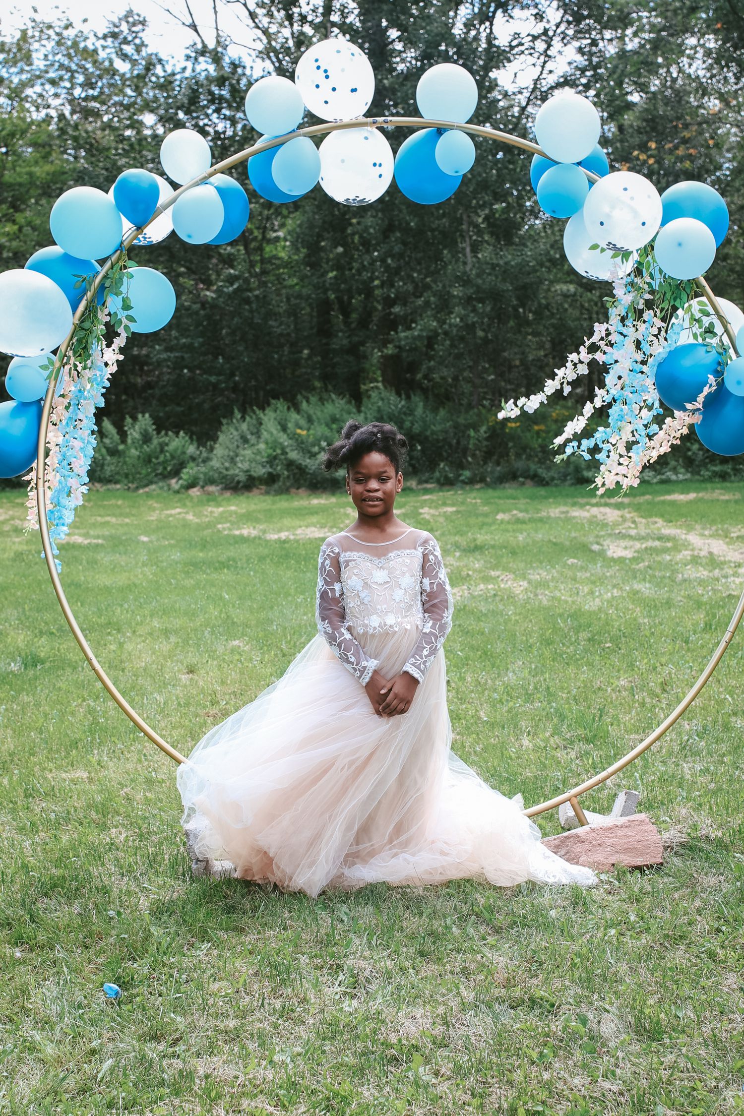 A bride in a flowing white lace dress poses gracefully on grass with blue and white balloons decorating the outdoor setting.