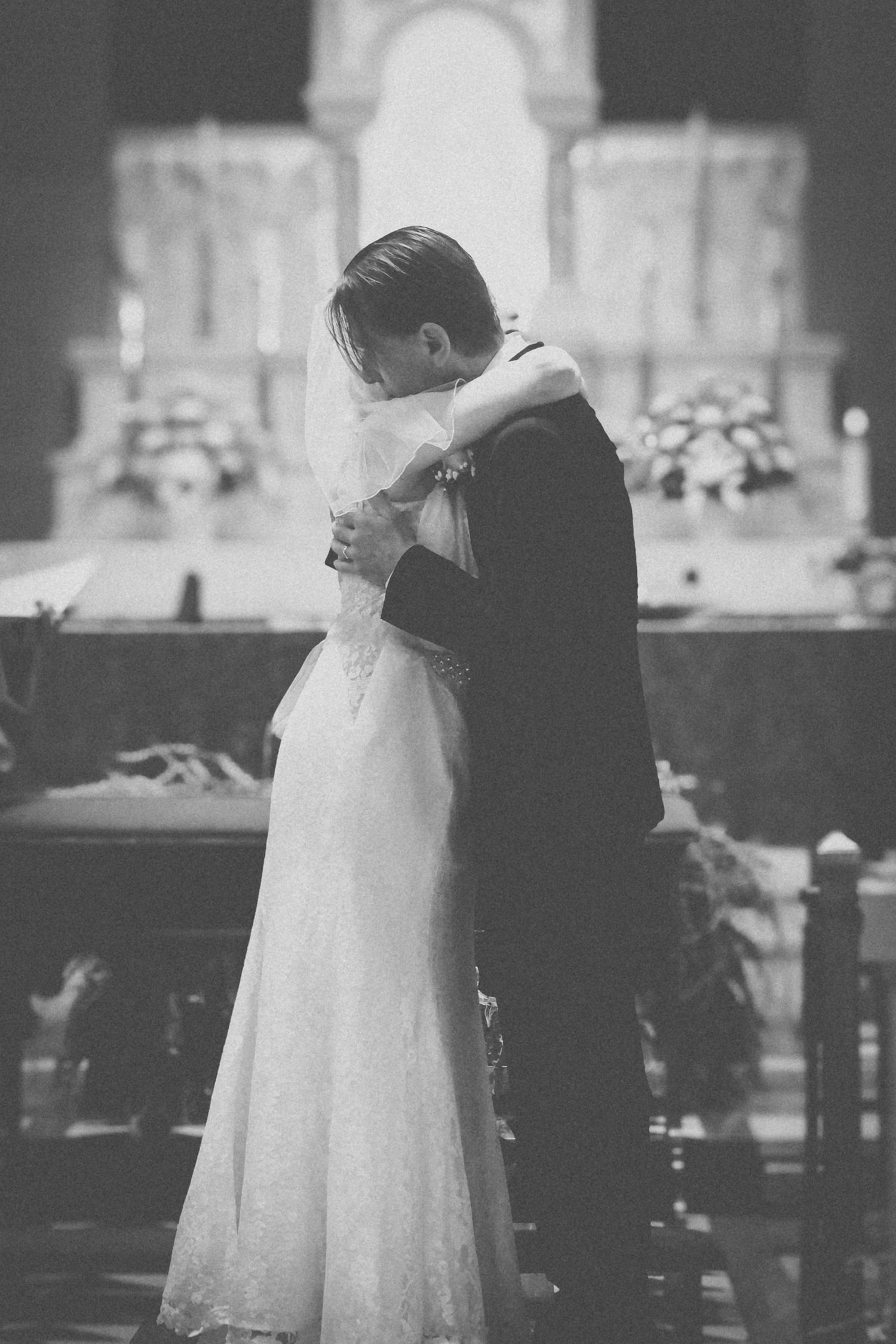 A couple embraces during their wedding ceremony in a church with ornate altar decorations and soft lighting.