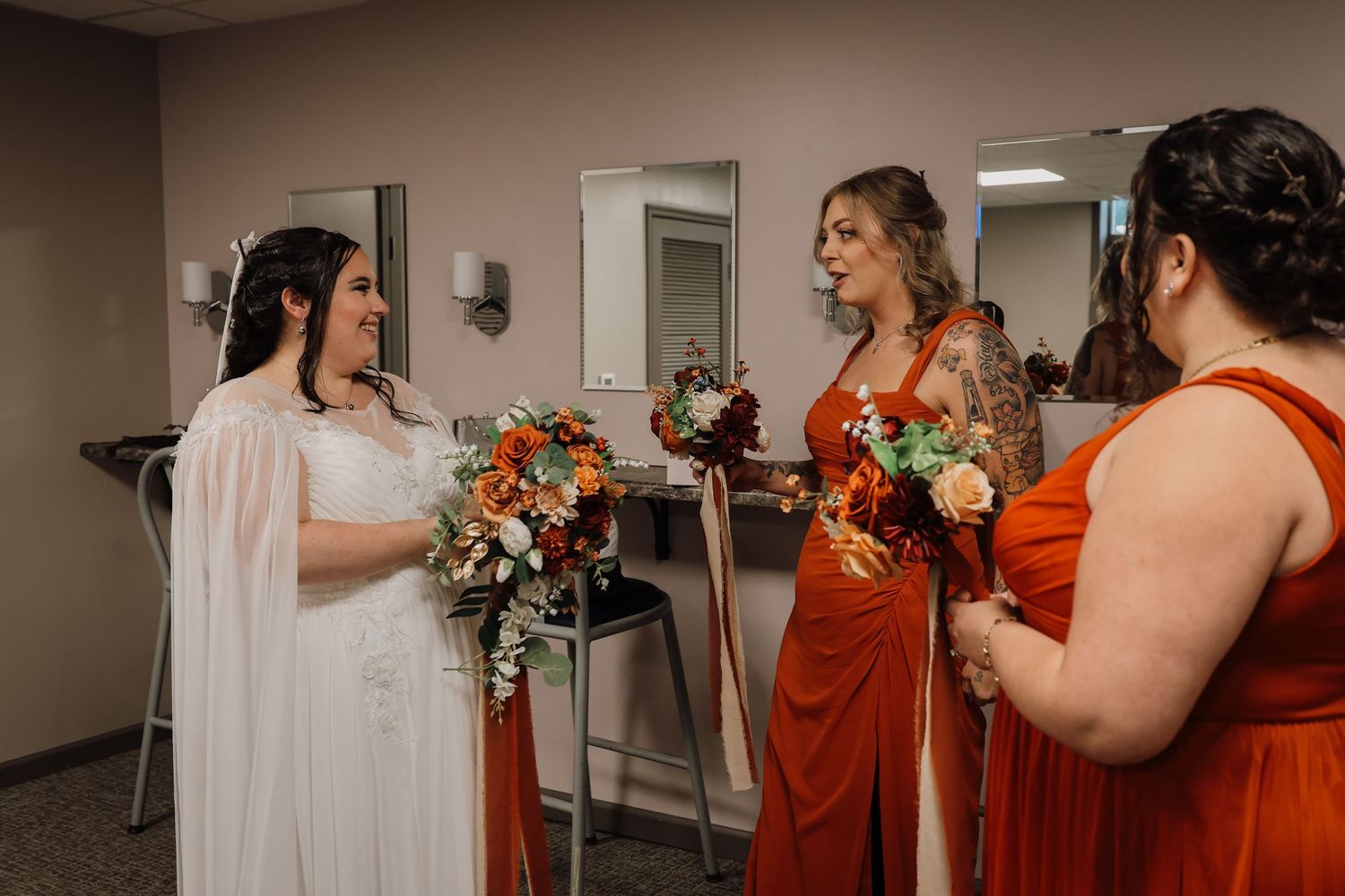 A group of bridesmaids in burnt orange dresses share an emotional moment with the bride in a white gown.