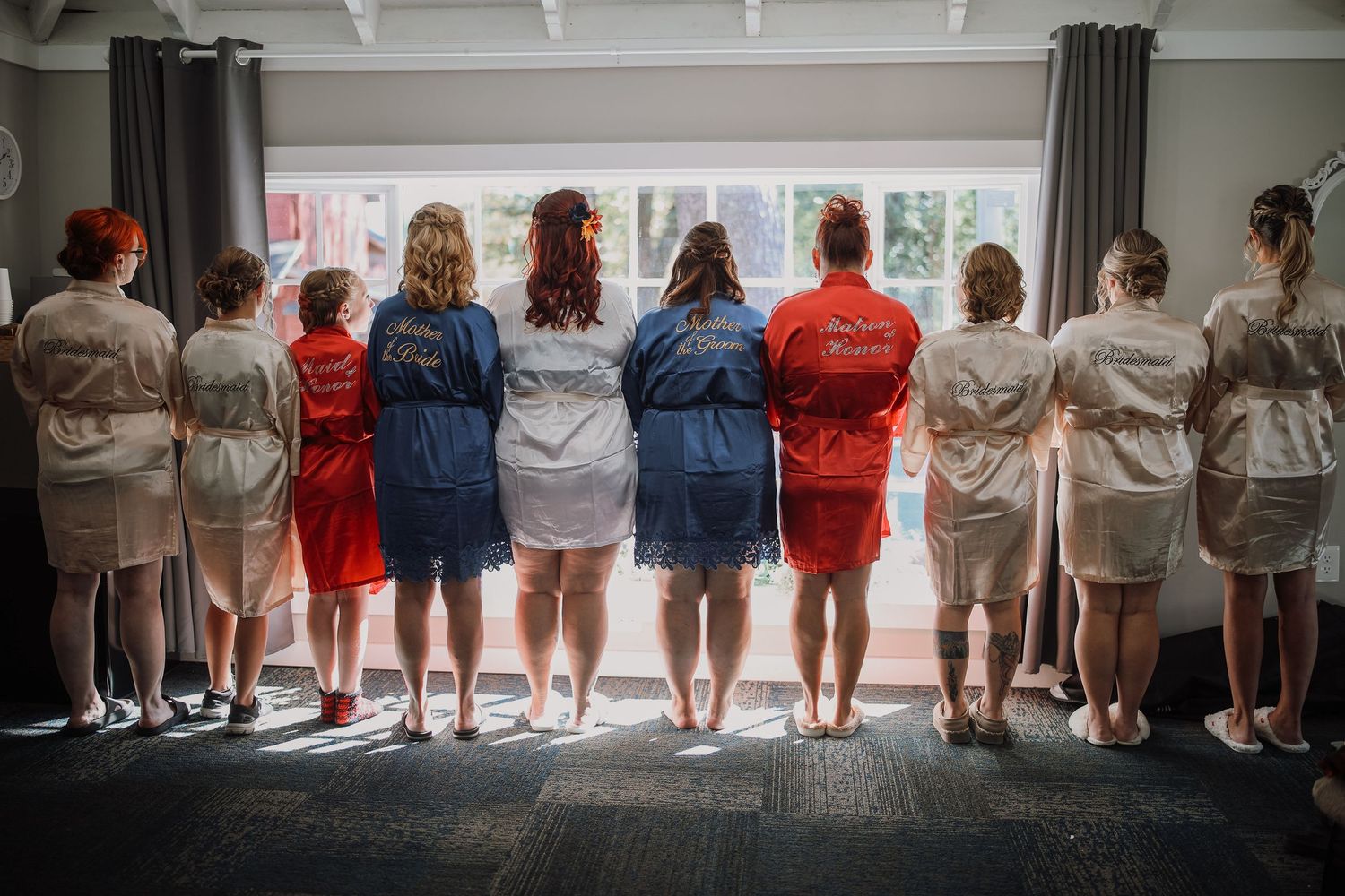 A group of people stand in a line facing a window, photographed from behind showing their backs and casual attire.