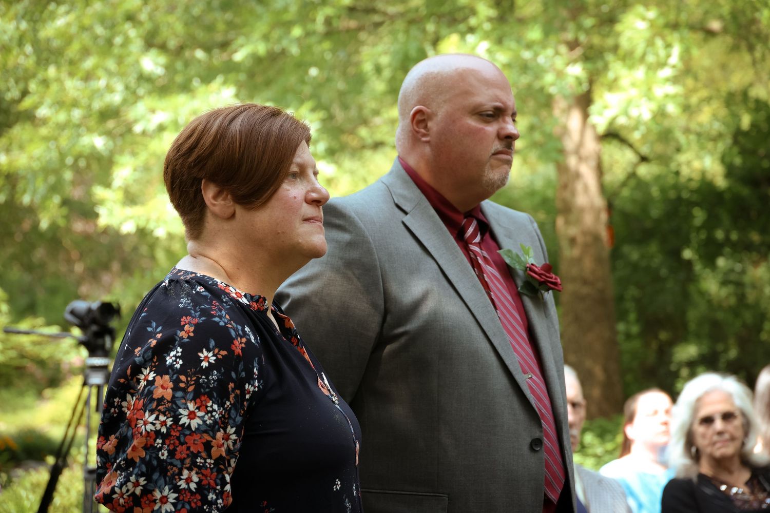 A couple in formal attire stands together outdoors during a garden event with sunlit trees in the background.