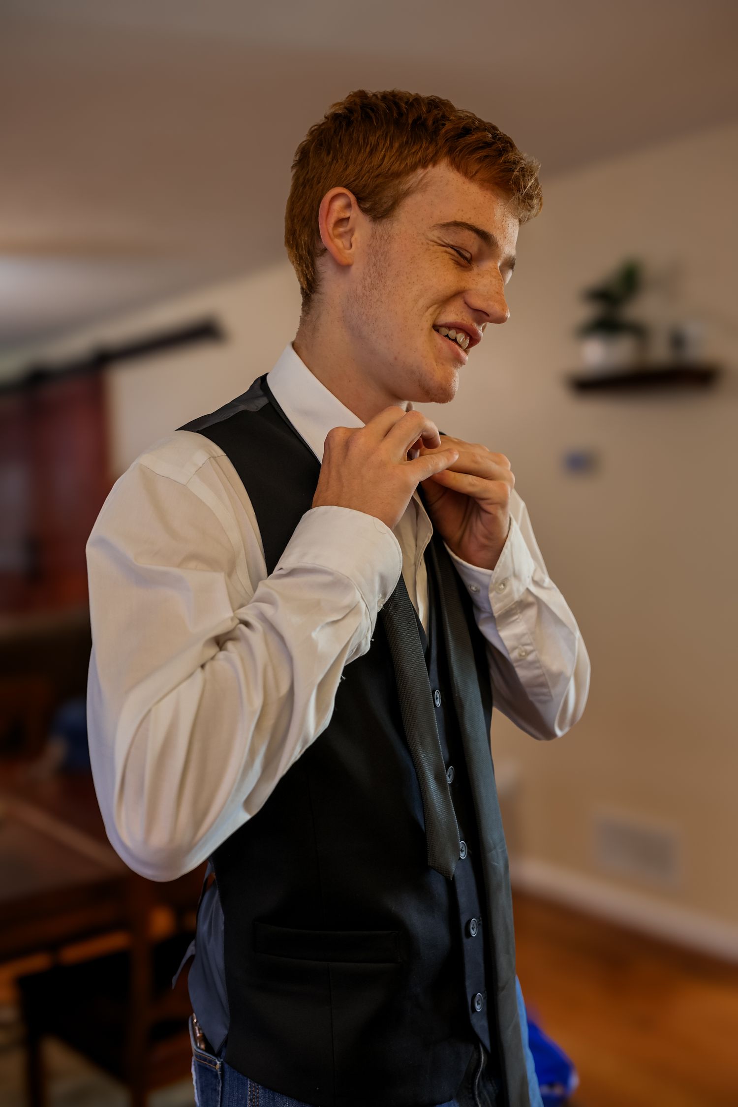 A person in formal attire adjusts their tie while smiling in a room with hardwood floors and indoor lighting.
