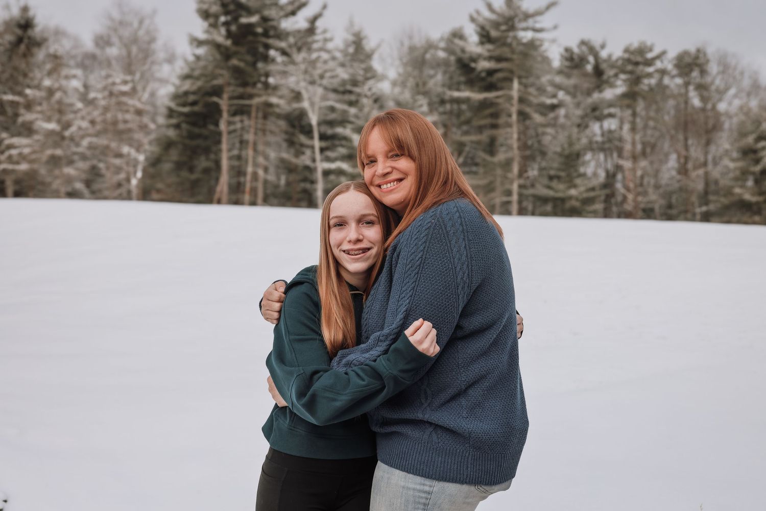 Two people embrace and laugh together in a snowy winter forest setting while wearing cozy sweaters.