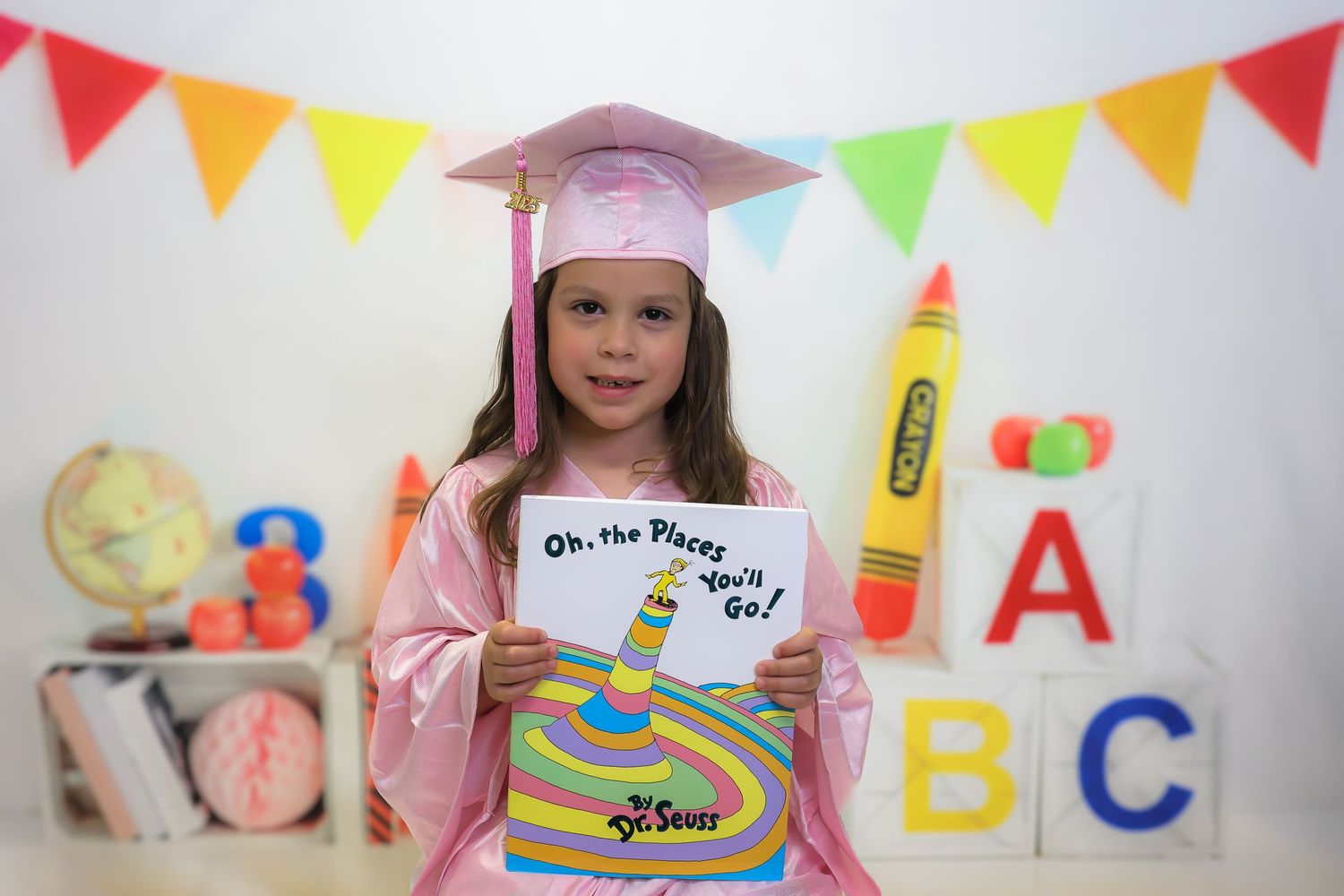 A student in pink graduation cap and gown holds up a Dr. Seuss book with ABC decorations and colorful bunting in background.