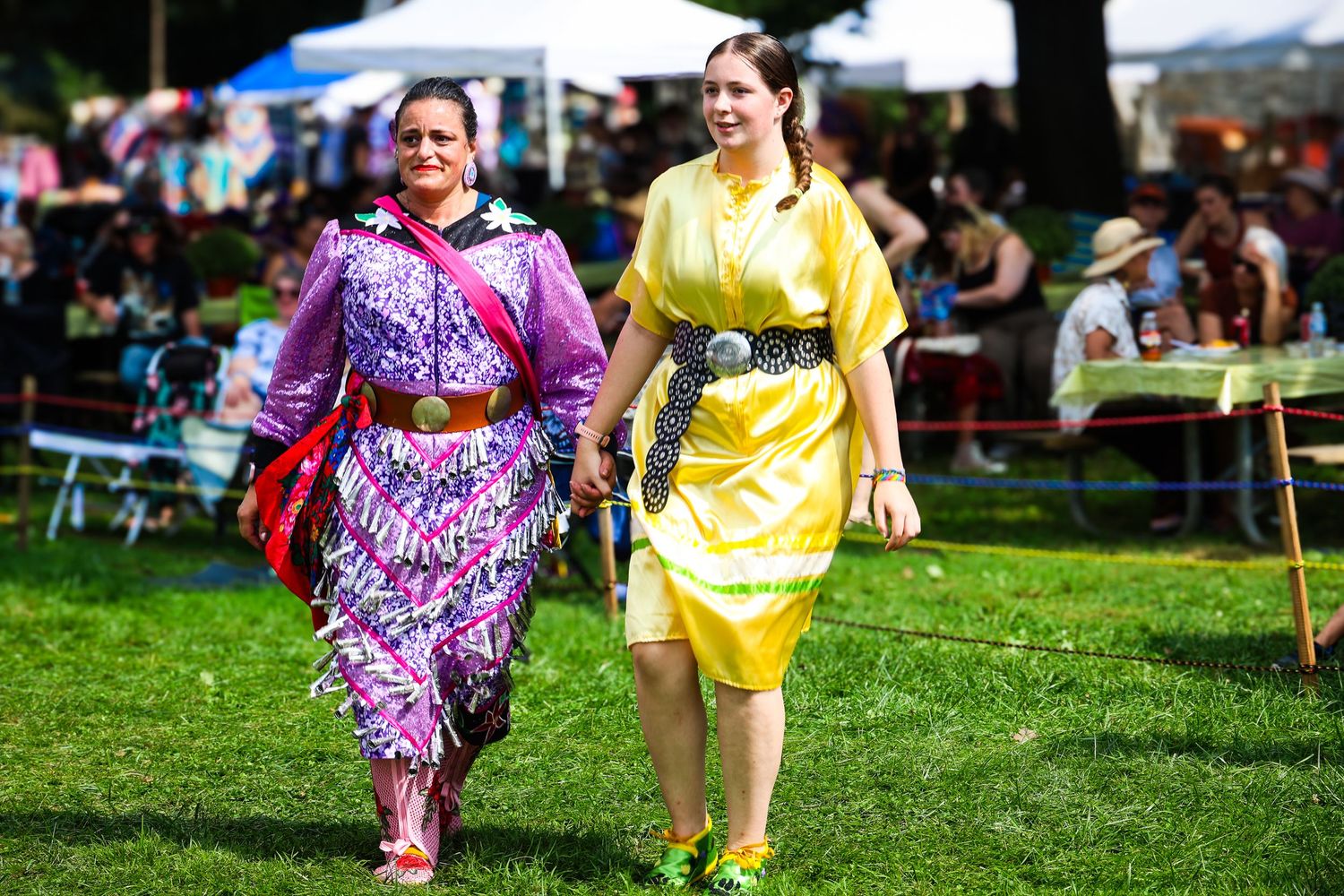 Two dancers in vibrant purple and yellow costumes perform together on a grassy lawn at an outdoor festival.