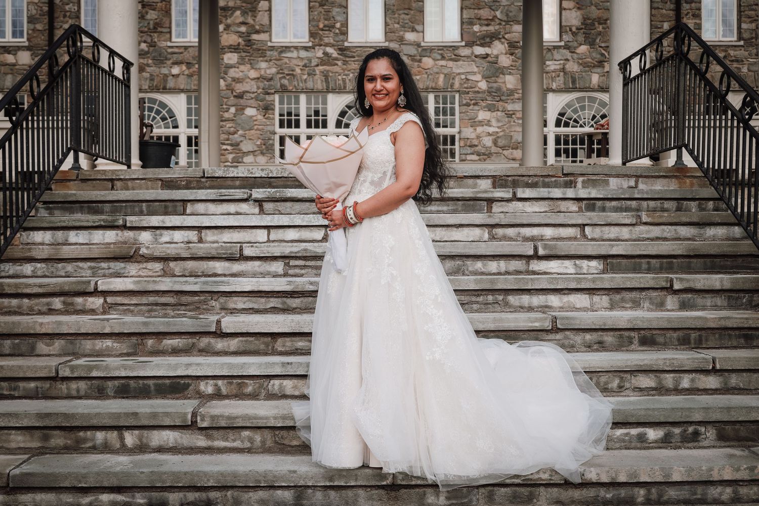 A bride in a white ballgown poses on grand stone steps of a historic building.