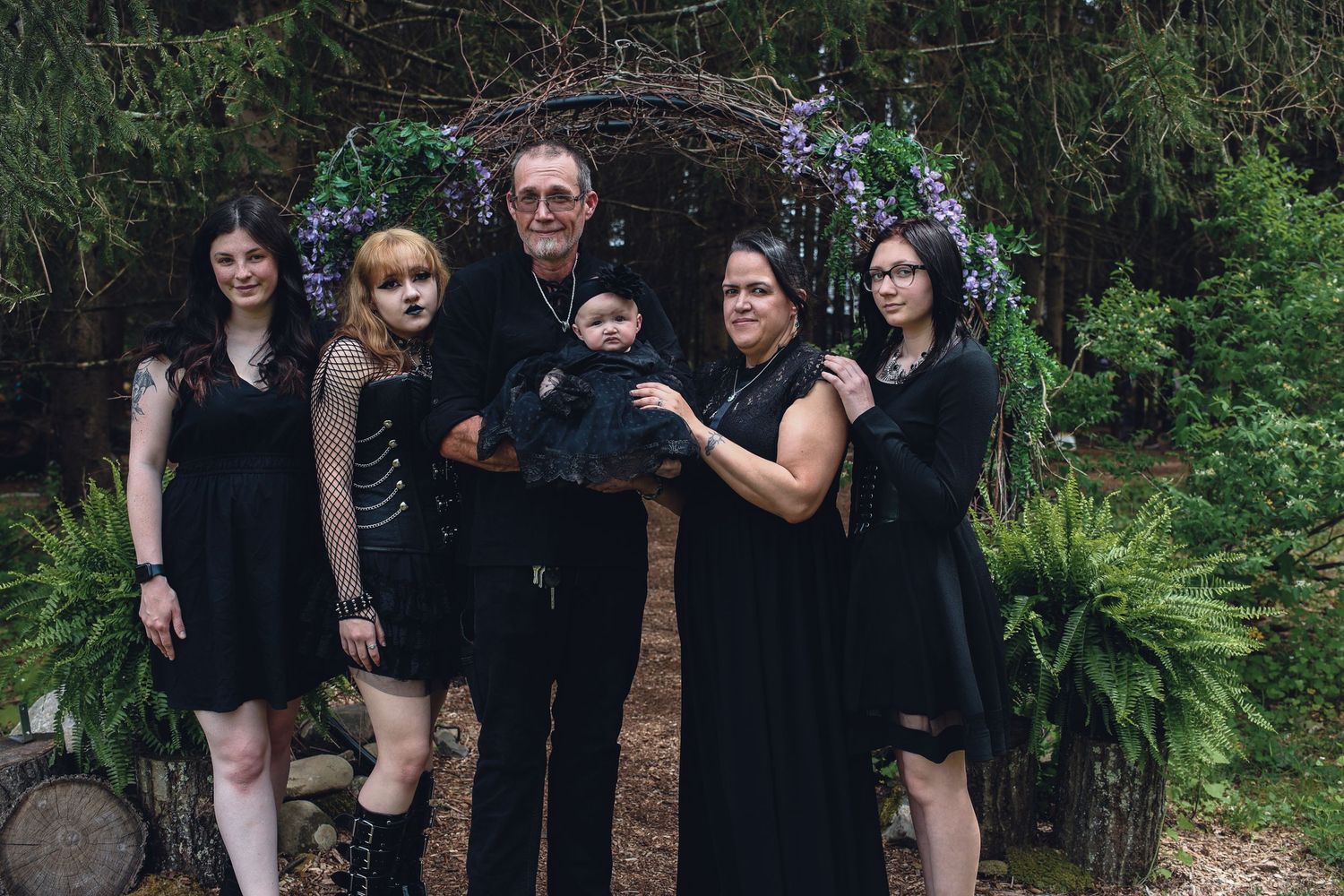A group wearing black outfits poses together in a lush garden setting during a photo session.
