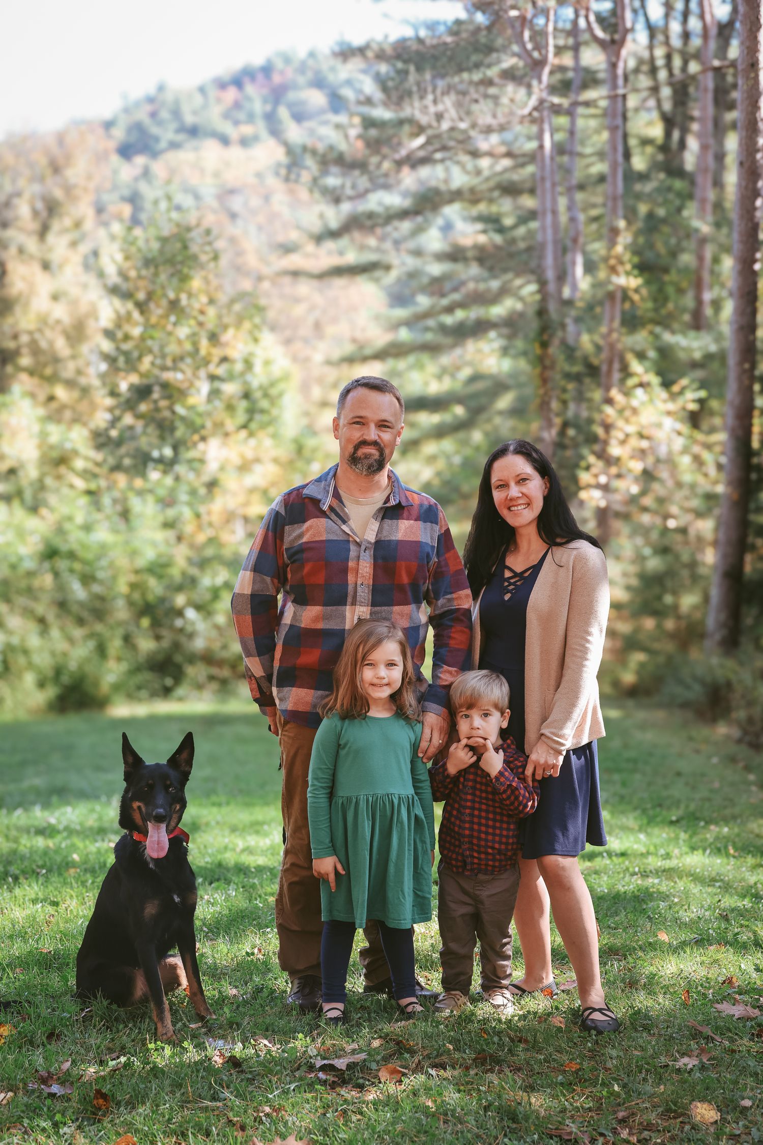 A family and their black dog pose together on a grassy hillside during autumn.