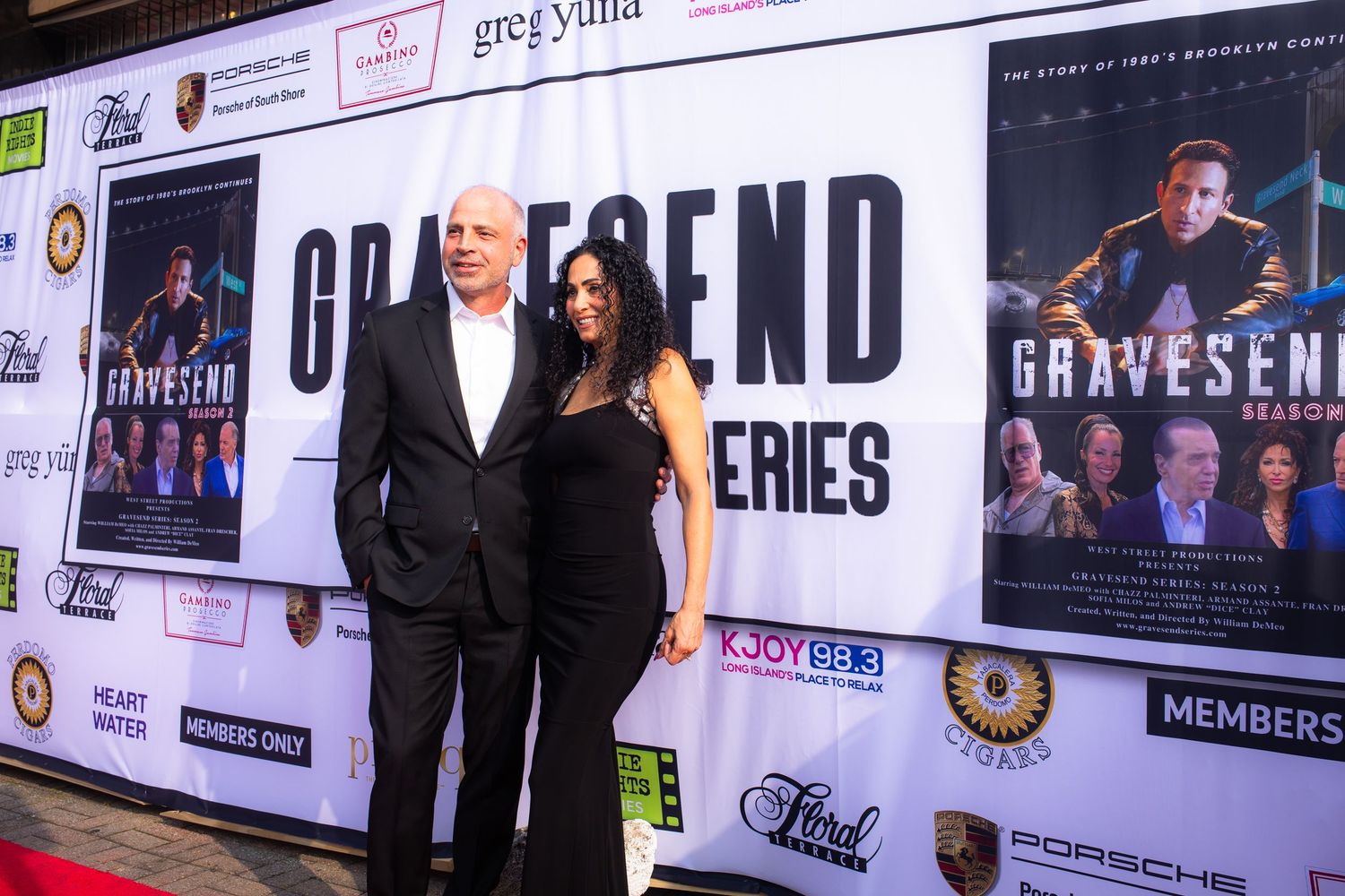 Event attendees pose in front of a backdrop covered with sponsor logos and promotional banners at a formal gathering.