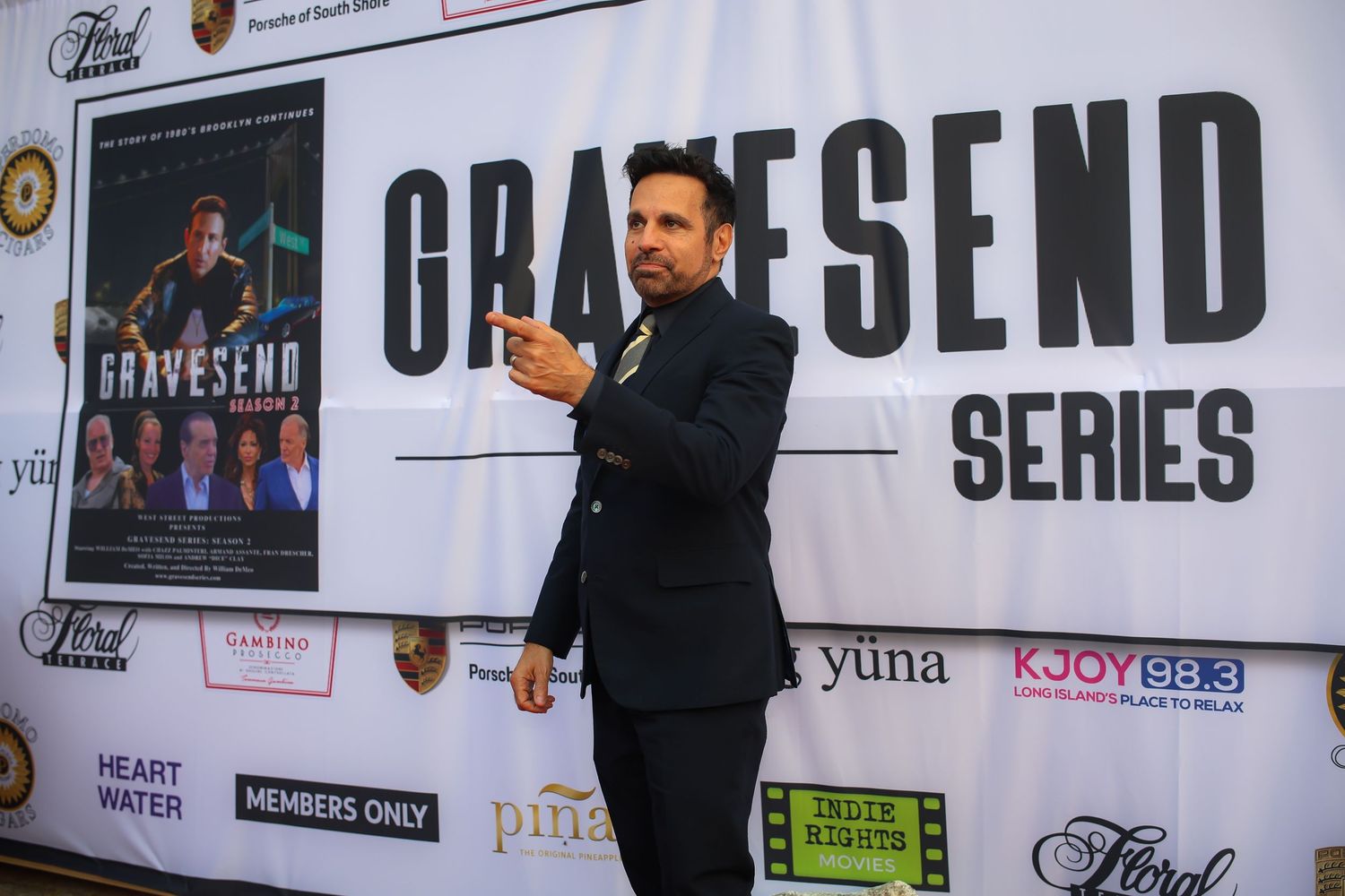 A person in a dark suit gestures while standing in front of a large banner display at an event series.