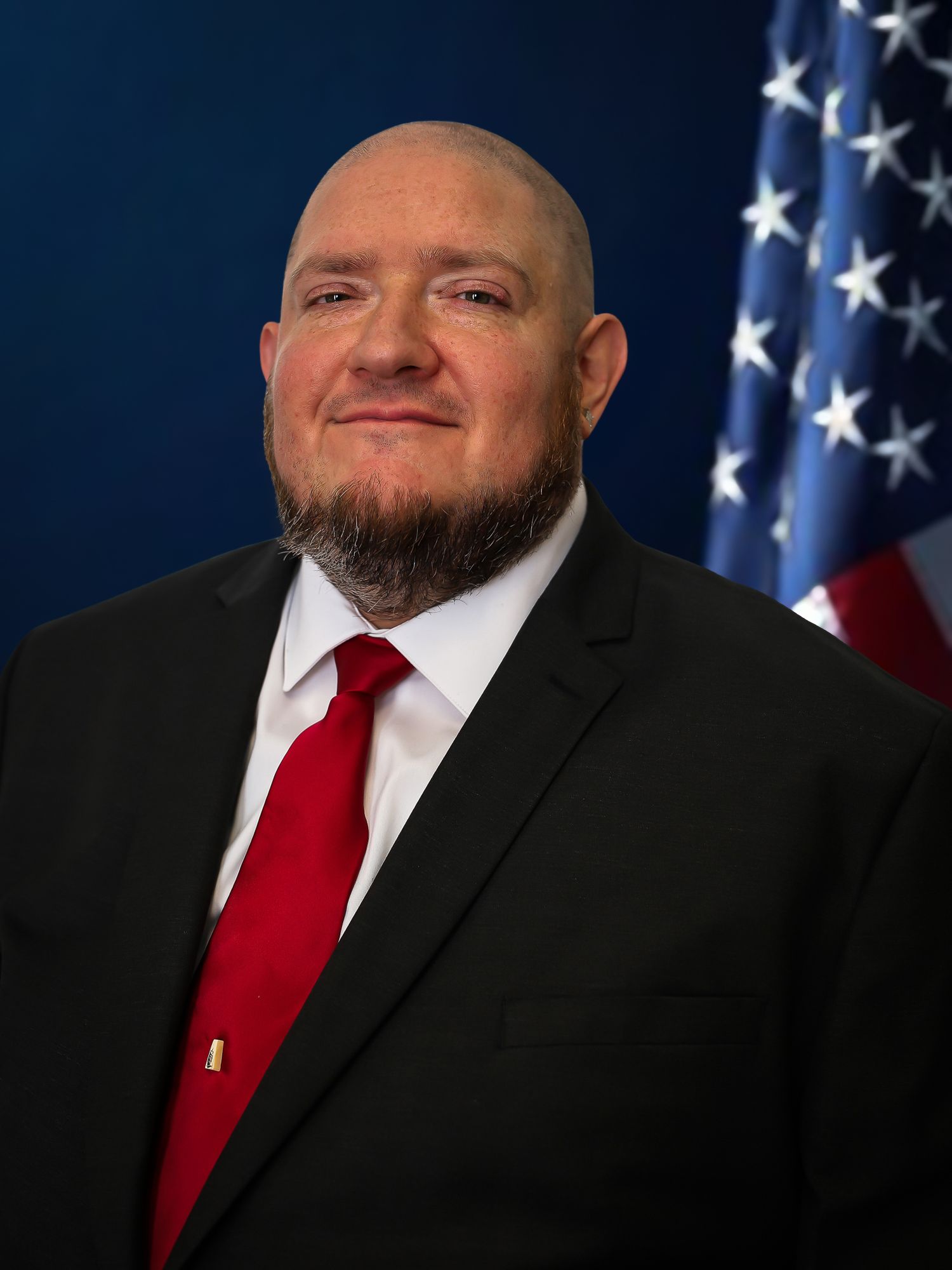 Professional headshot portrait of a person in a black suit and red tie standing in front of an American flag.