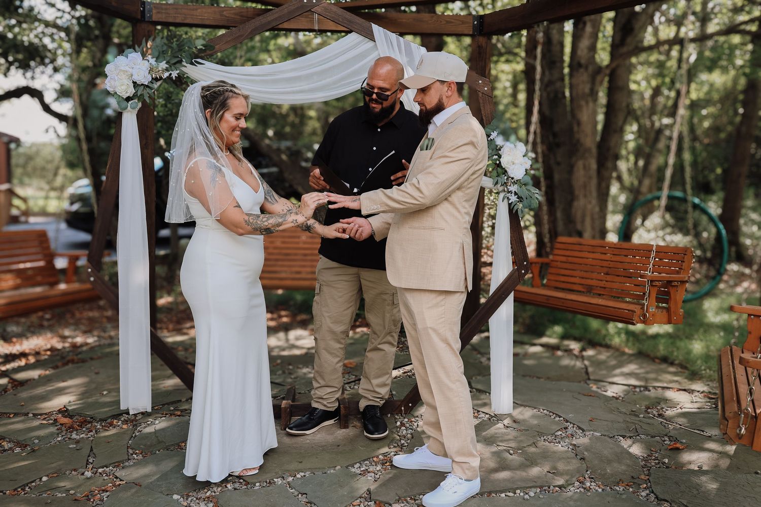 A couple exchanges rings during an outdoor wedding ceremony under a rustic wooden pergola.