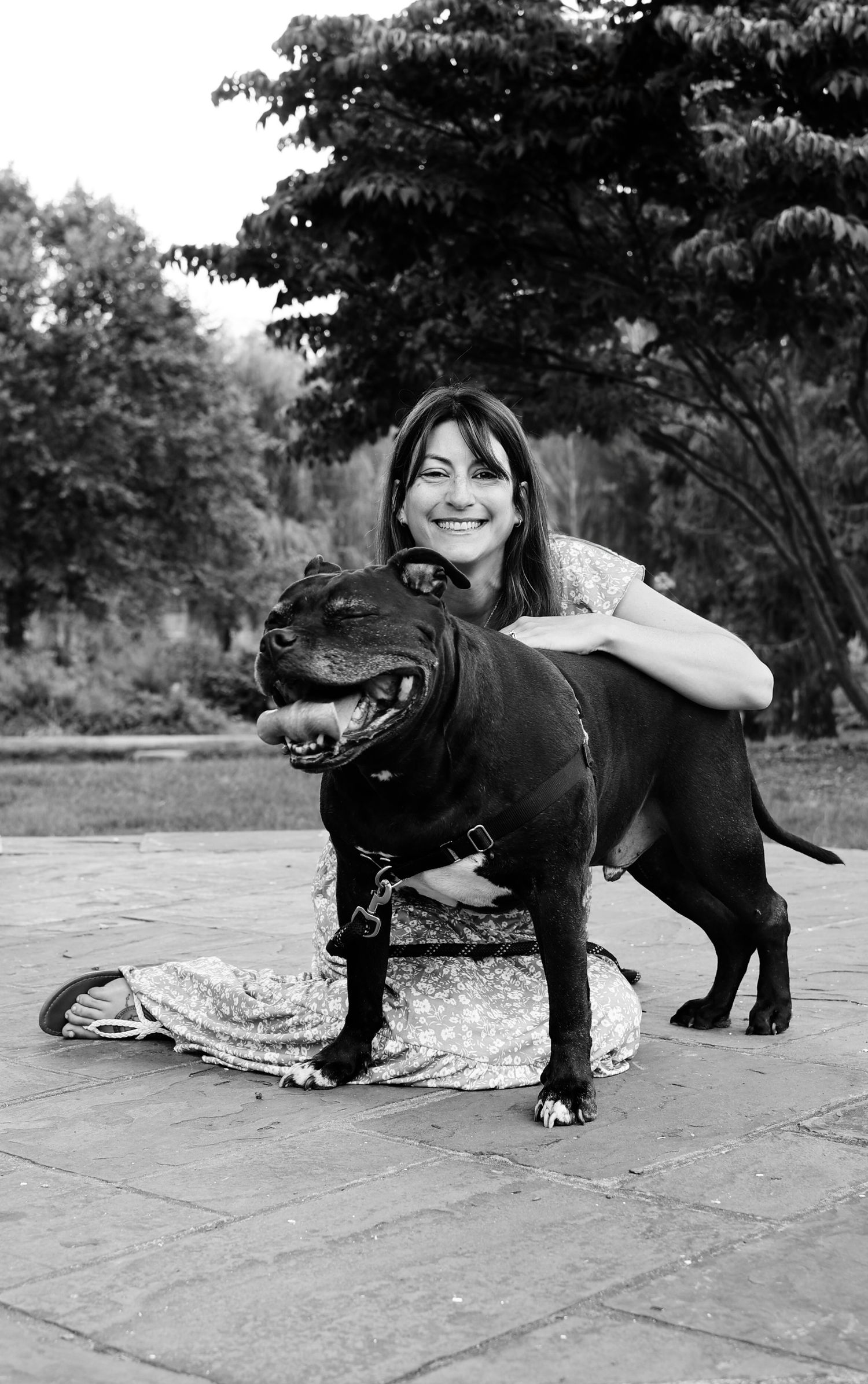Black and white portrait of a person posing with a happy black Staffordshire Bull Terrier dog on a stone patio.