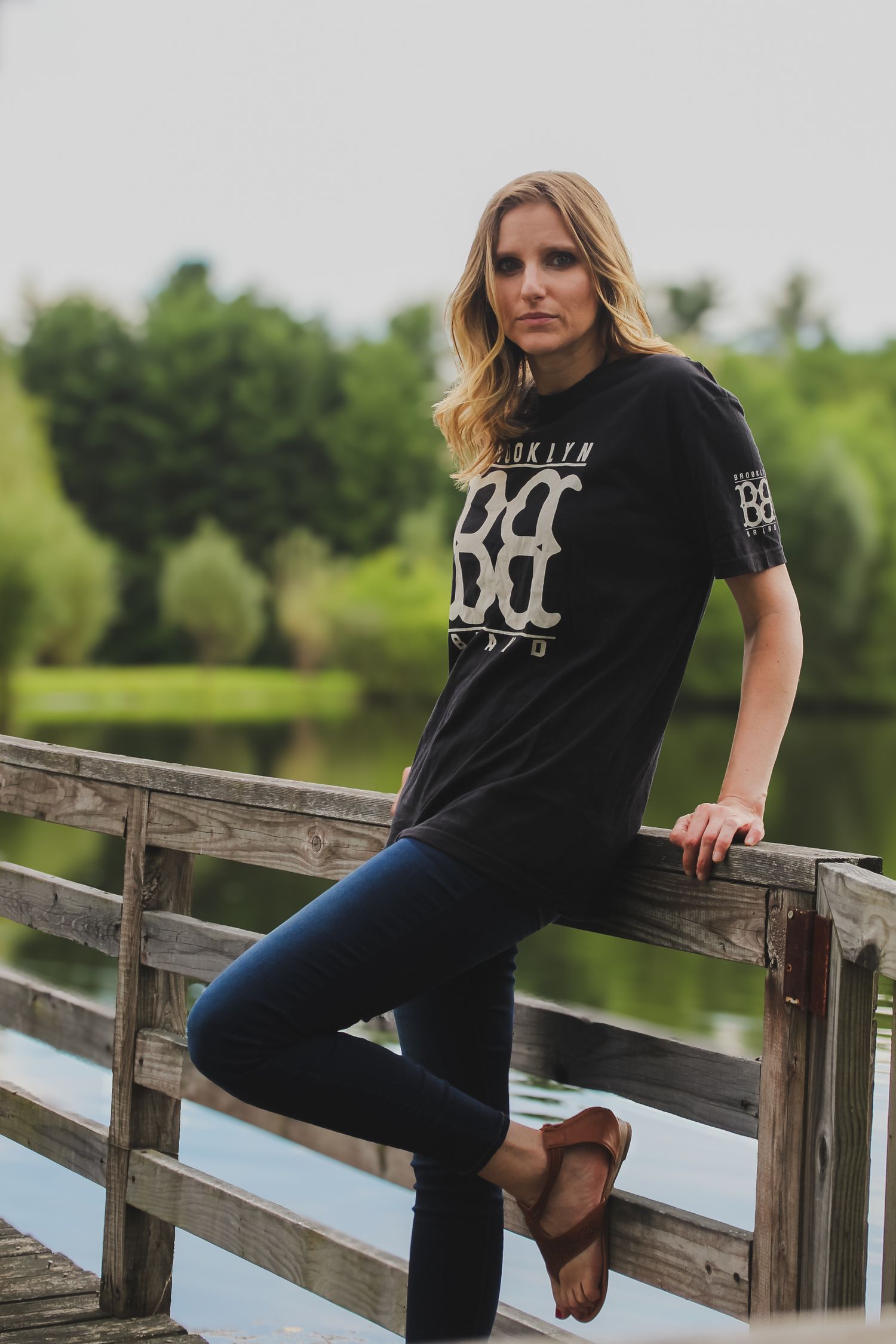Casual outdoor portrait of someone wearing a black graphic t-shirt and jeans while leaning on wooden railing by a lake.