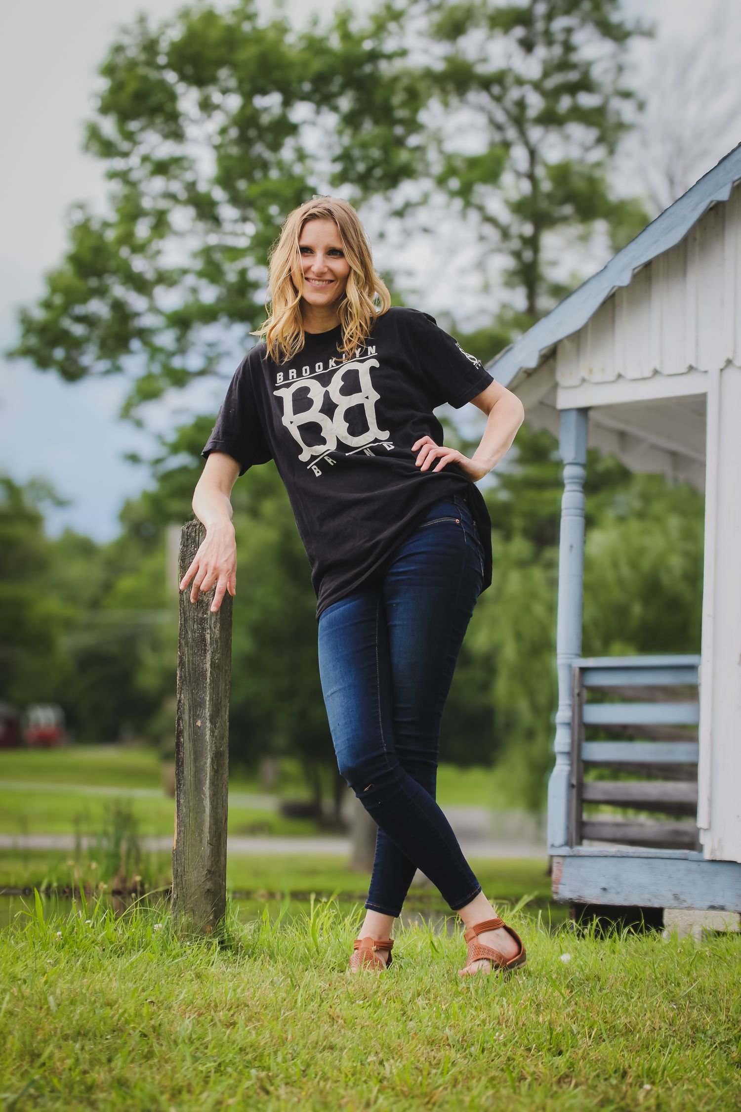A person in a black t-shirt and jeans leans casually against a wooden fence post in a rural country setting.