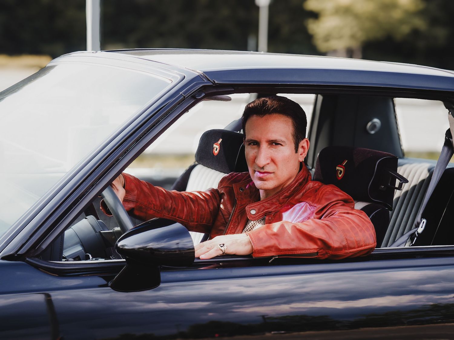 A person in a reddish leather jacket sits behind the steering wheel of a black vehicle.