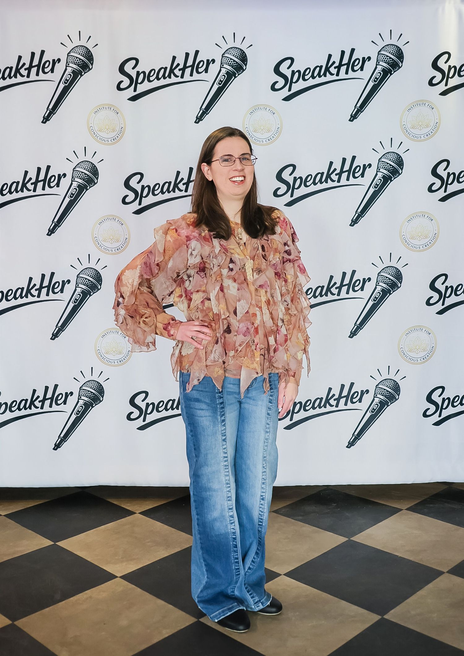 Woman in floral blouse and jeans posing at SpeakHer event backdrop with microphone logos.