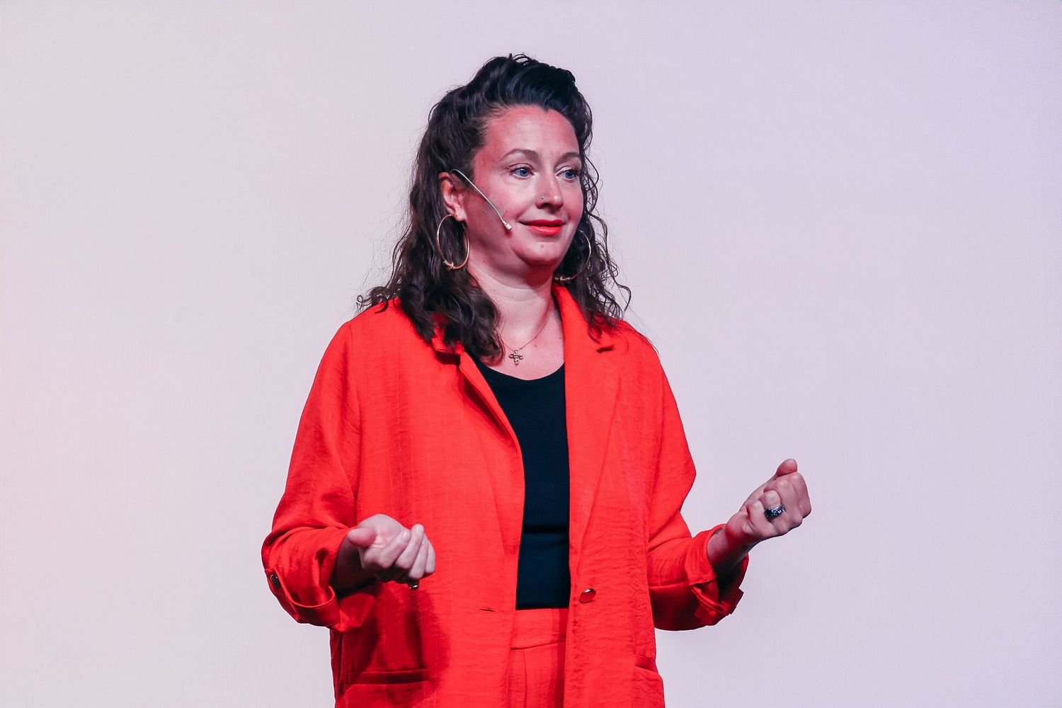 Woman in red blazer speaking on stage with a microphone, gesturing confidently during a presentation.
