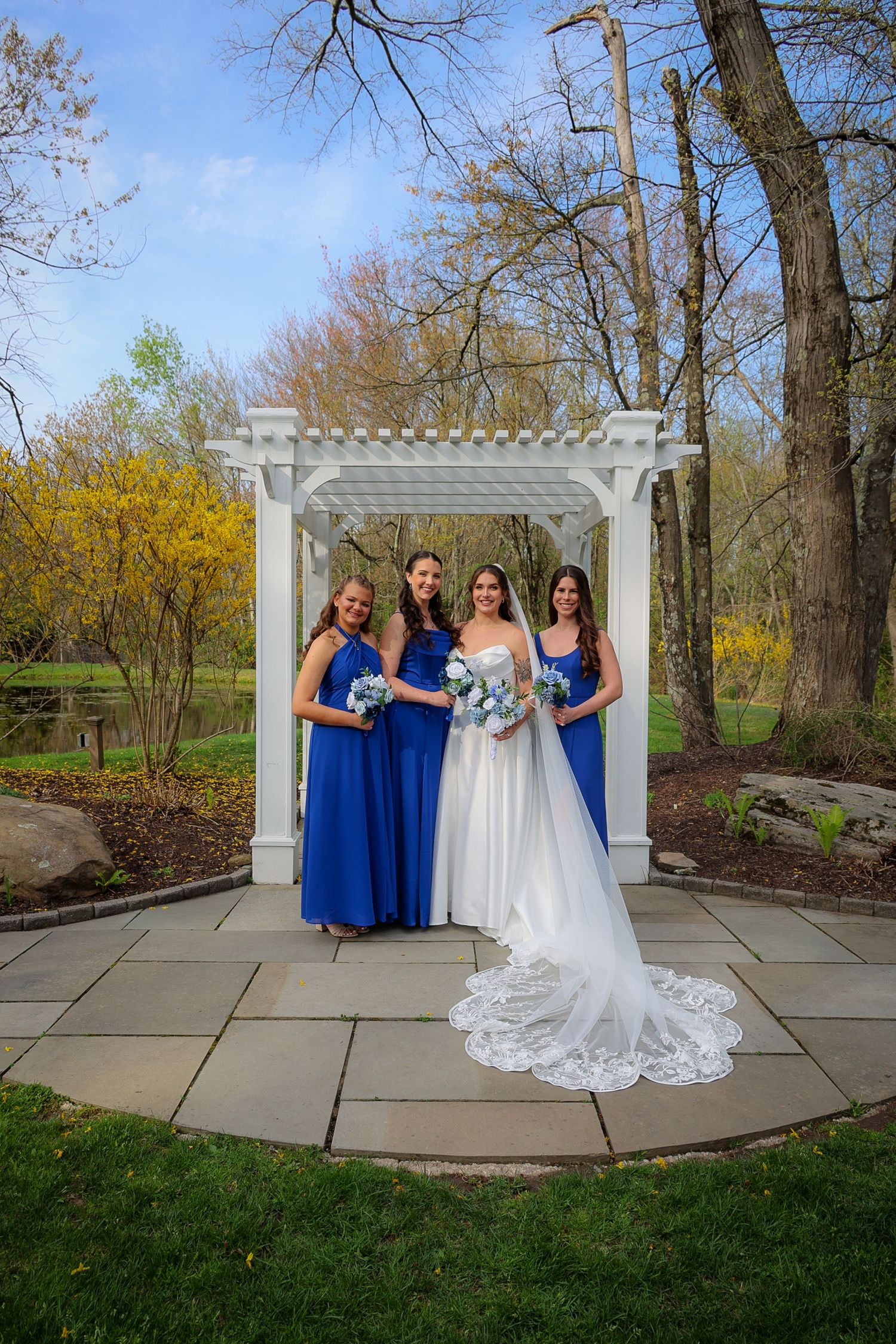 Bride in white gown with two bridesmaids in royal blue dresses holding bouquets under white pergola outdoors.
