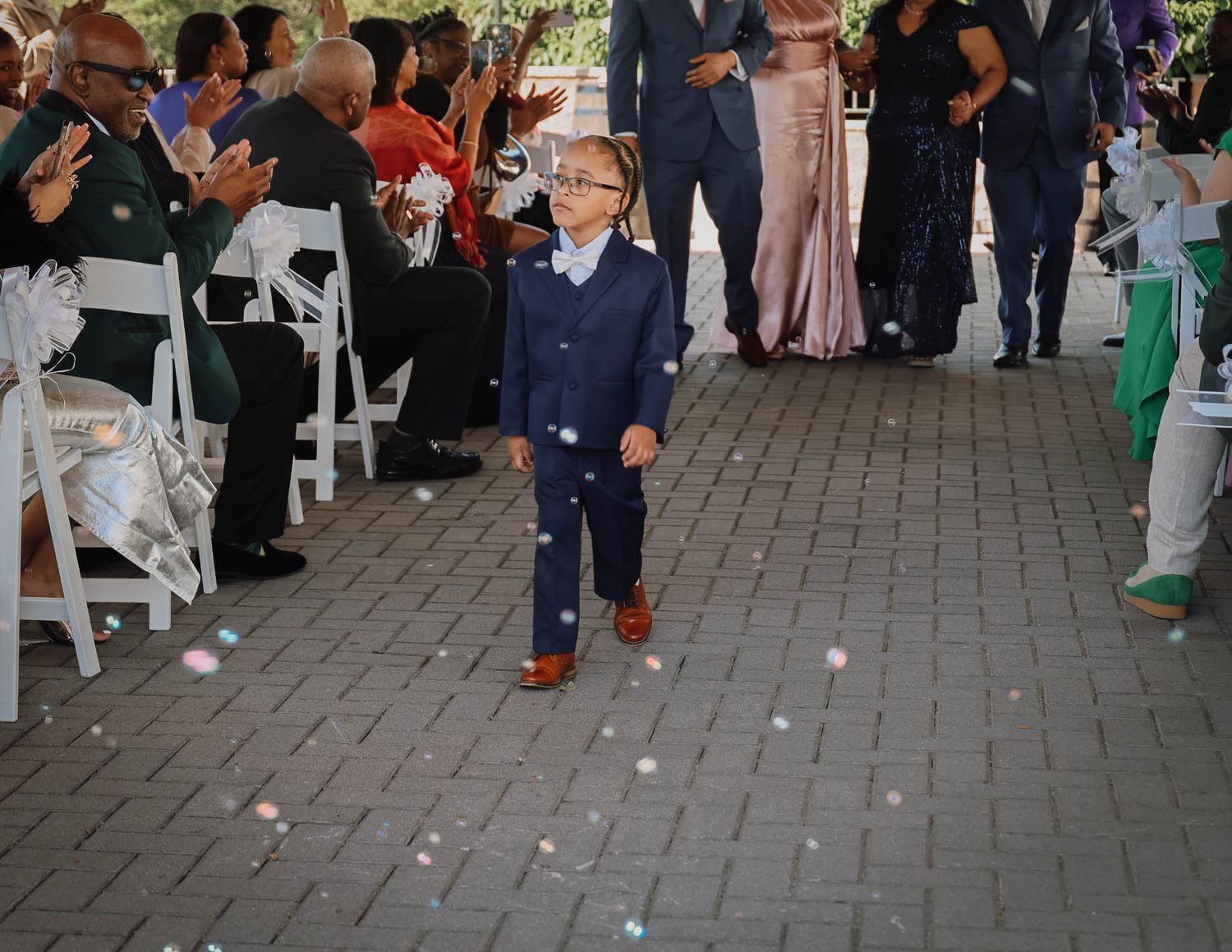 Young boy in navy suit walks down wedding aisle as flower petals fall around him at outdoor ceremony.