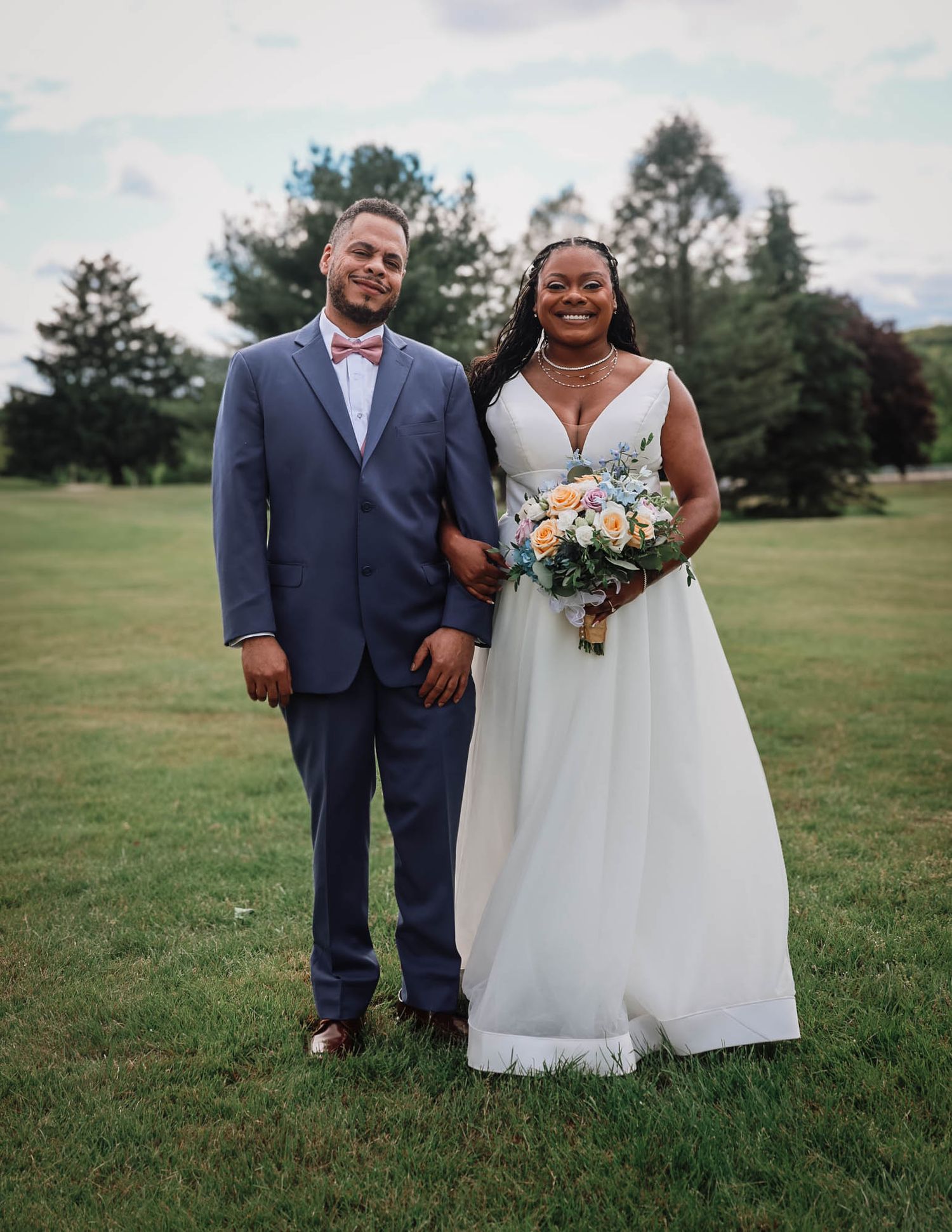 A couple poses outdoors on green grass, groom in blue suit with bow tie, bride in white gown holding a floral bouquet.