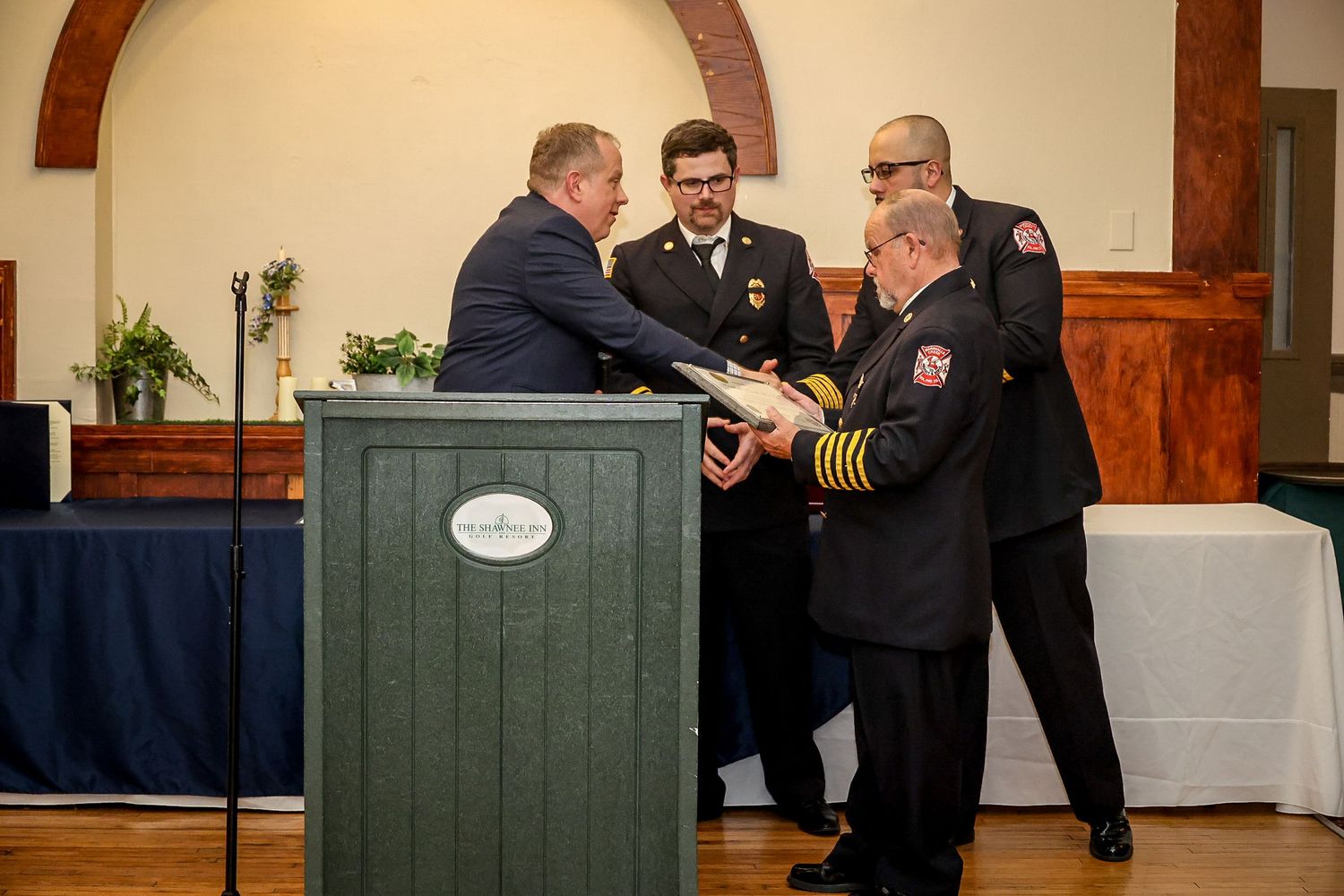 Firefighters in dress uniforms receive an award at a formal ceremony near a podium indoors.