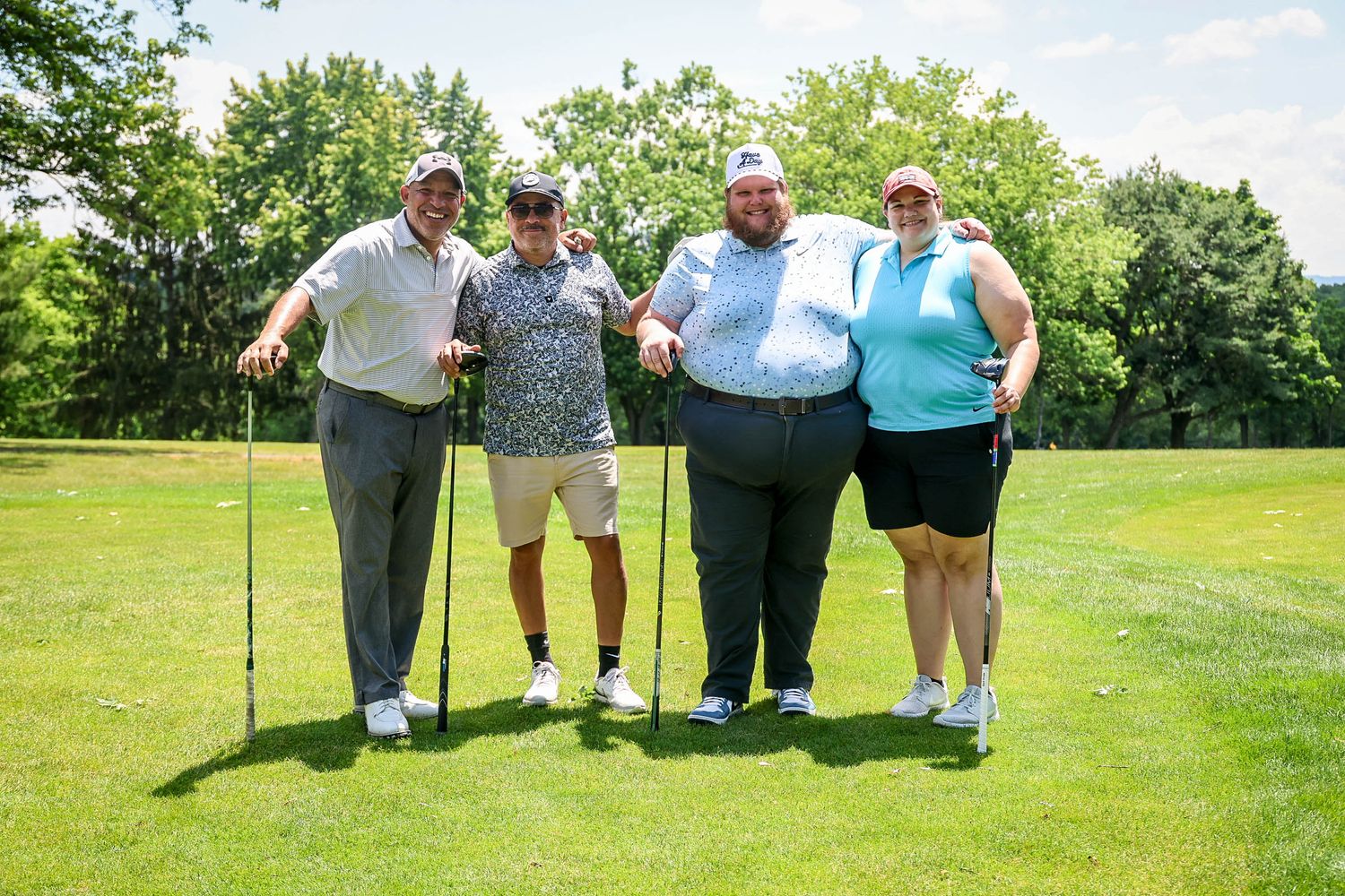 Four golfers posing together on a sunny green golf course, smiling and holding golf clubs.