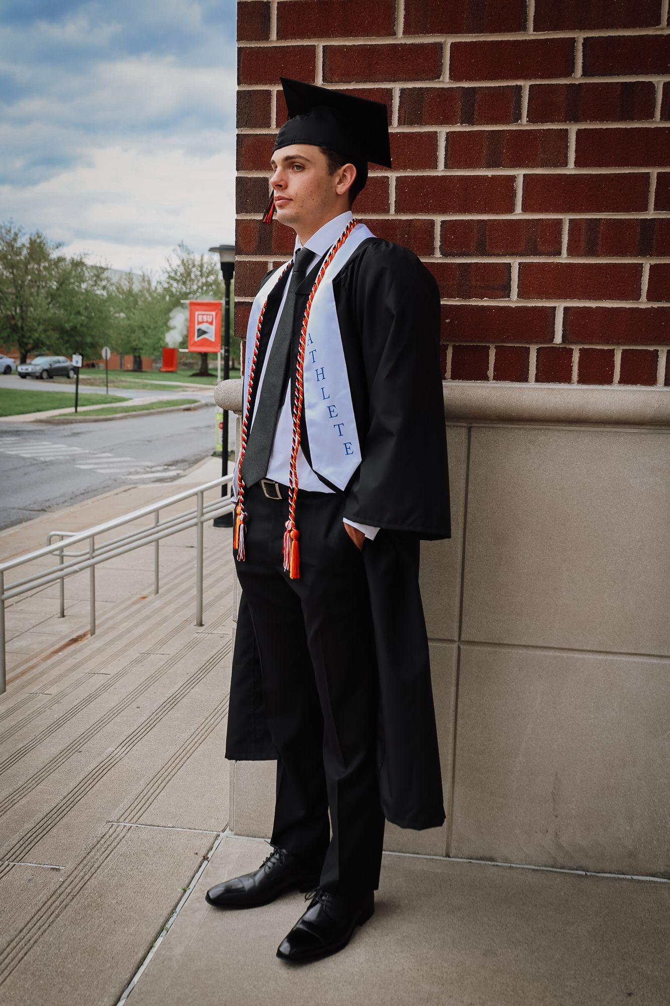 Young man in black graduation gown and cap with honor stole stands proudly outside brick building on graduation day.