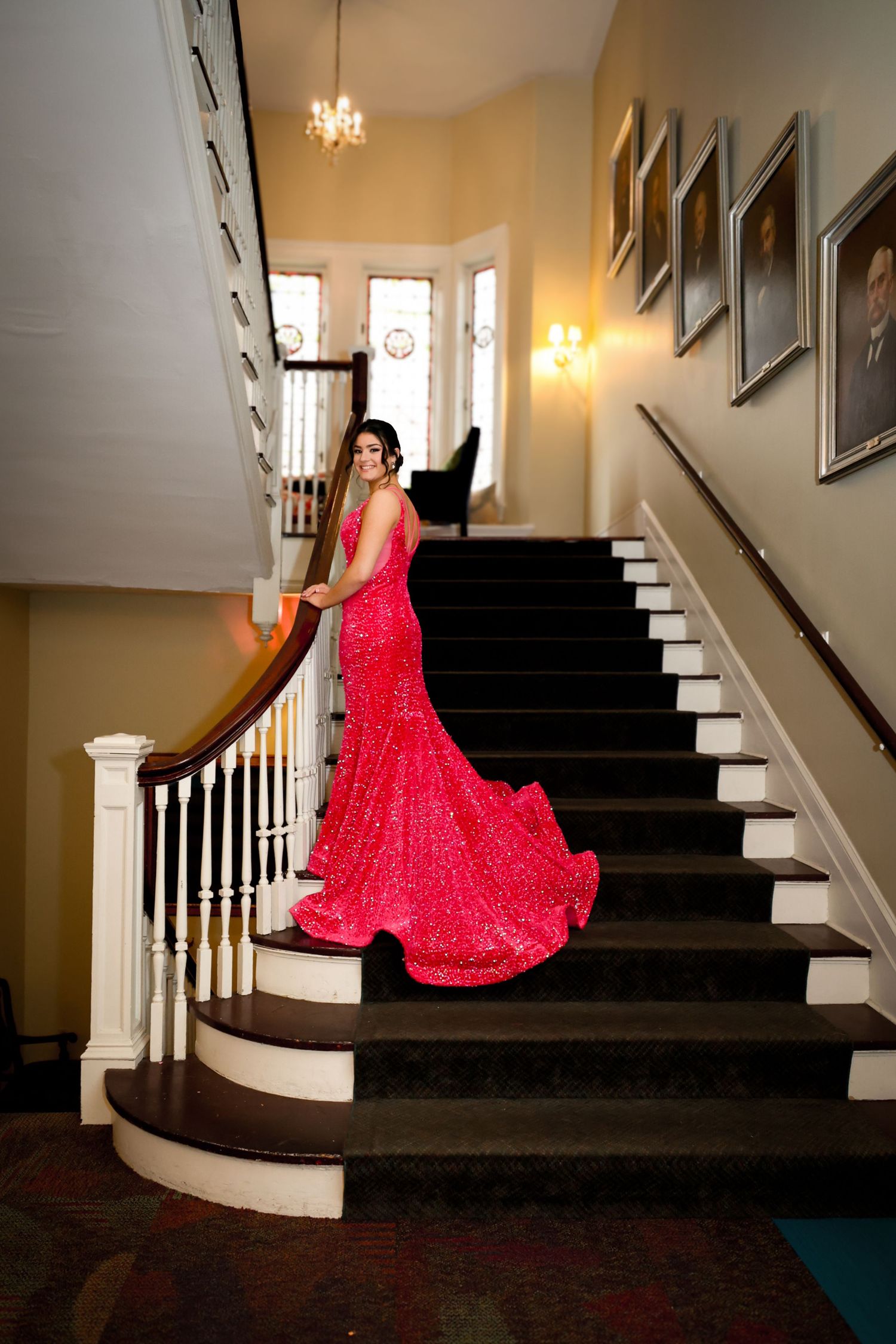 Woman in red sequin gown with train poses elegantly on grand staircase in elegant venue.
