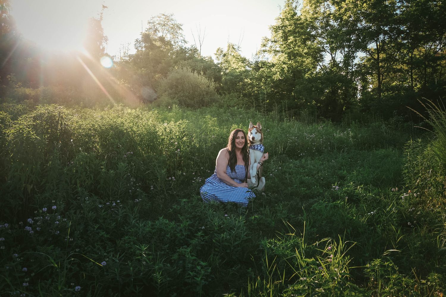 Woman in blue dress kneeling with a Husky dog in a sunlit green meadow surrounded by lush trees.