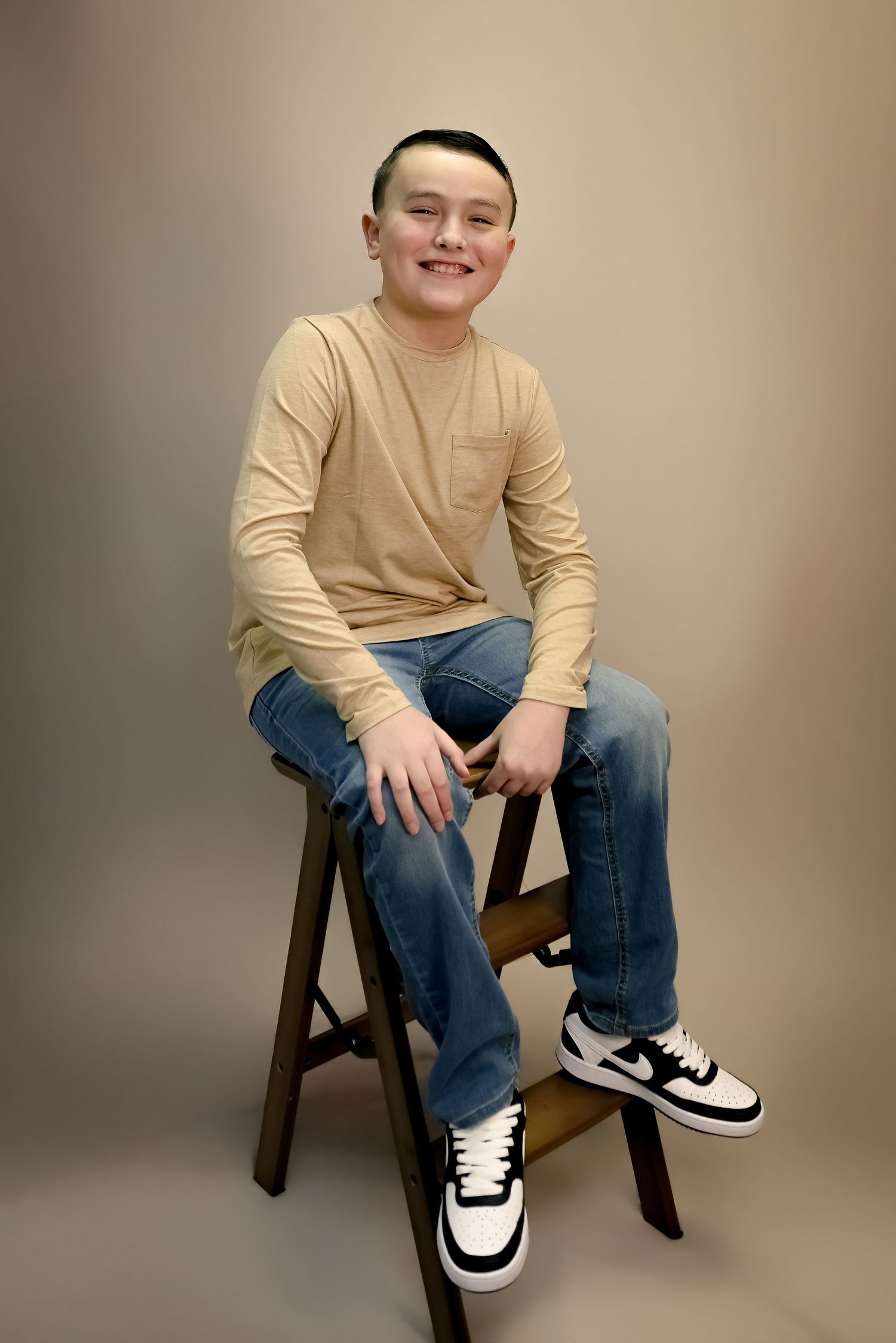 Smiling teen boy sits on wooden stool wearing beige long sleeve shirt, blue jeans, and black white Nike sneakers.