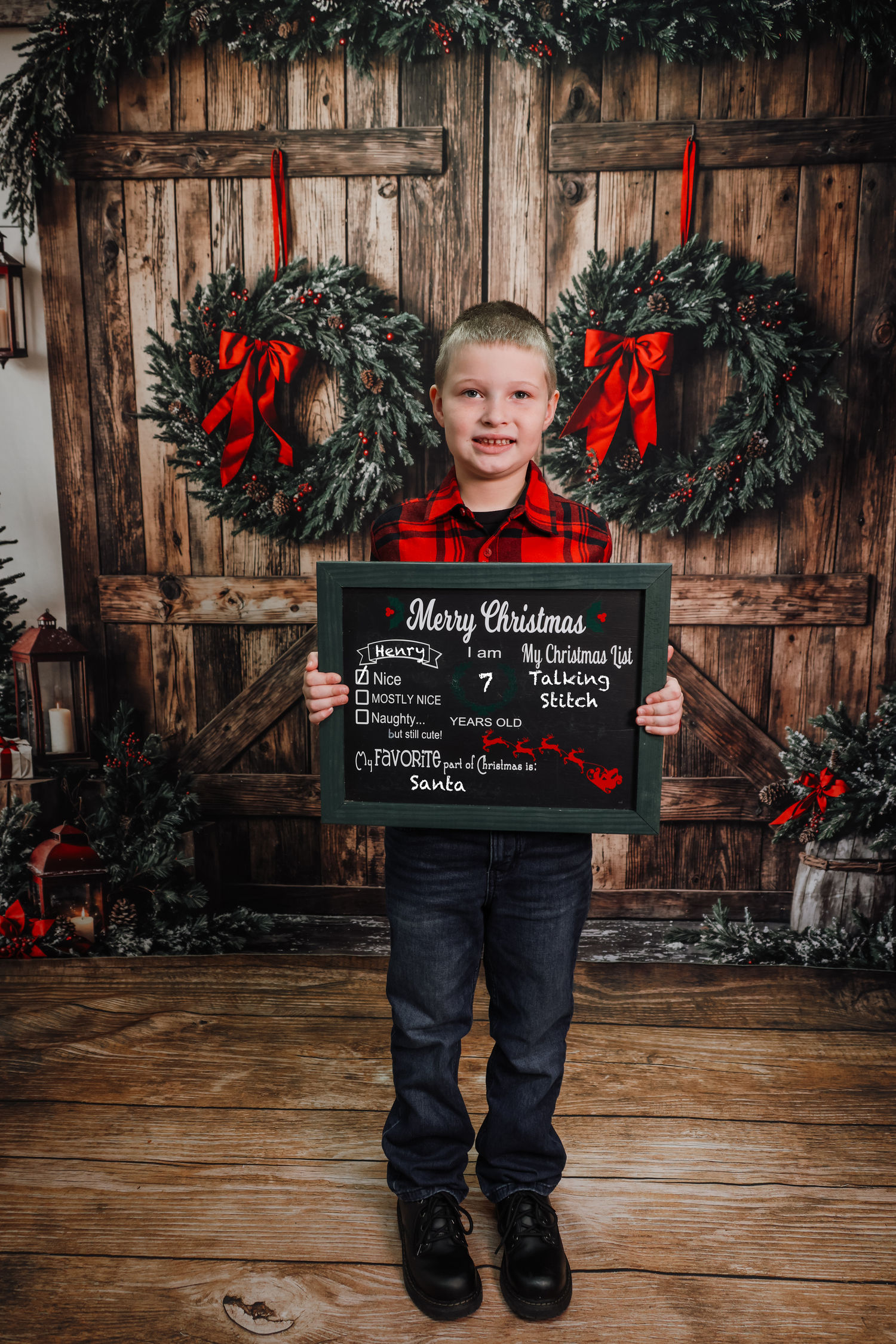 A young boy holds a Merry Christmas chalkboard sign, standing before rustic wooden doors adorned with festive wreaths.