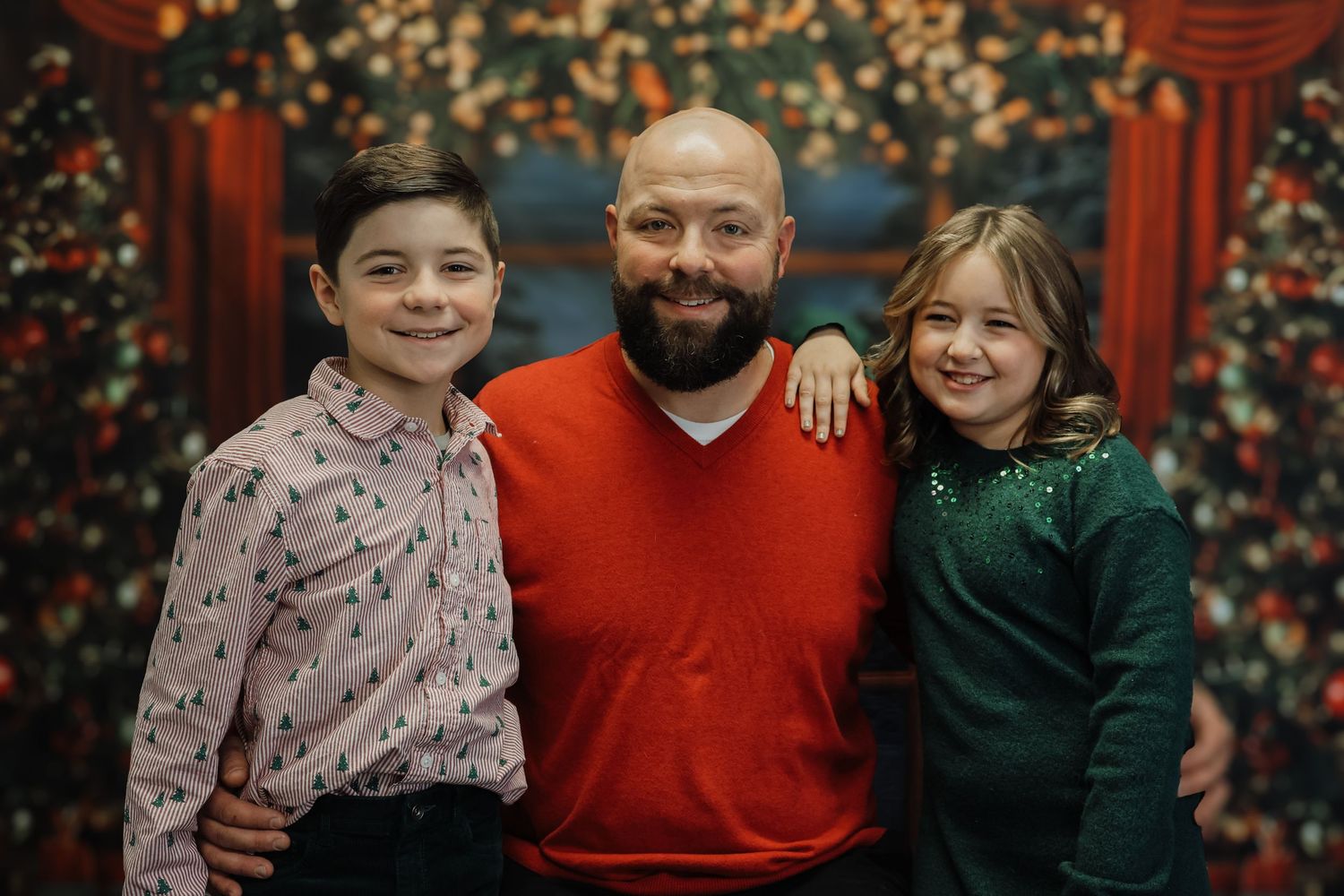A man in a red sweater poses with two children in front of a festive Christmas backdrop for a holiday photo.