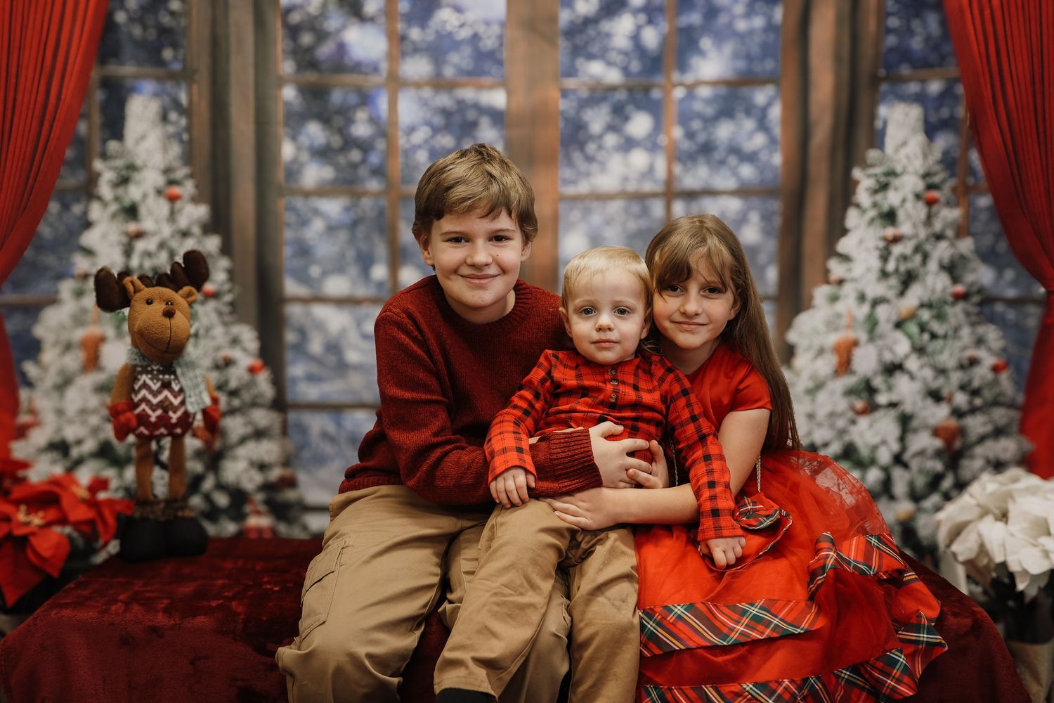 Three children pose together in festive holiday attire before decorated Christmas trees and snowy windows.