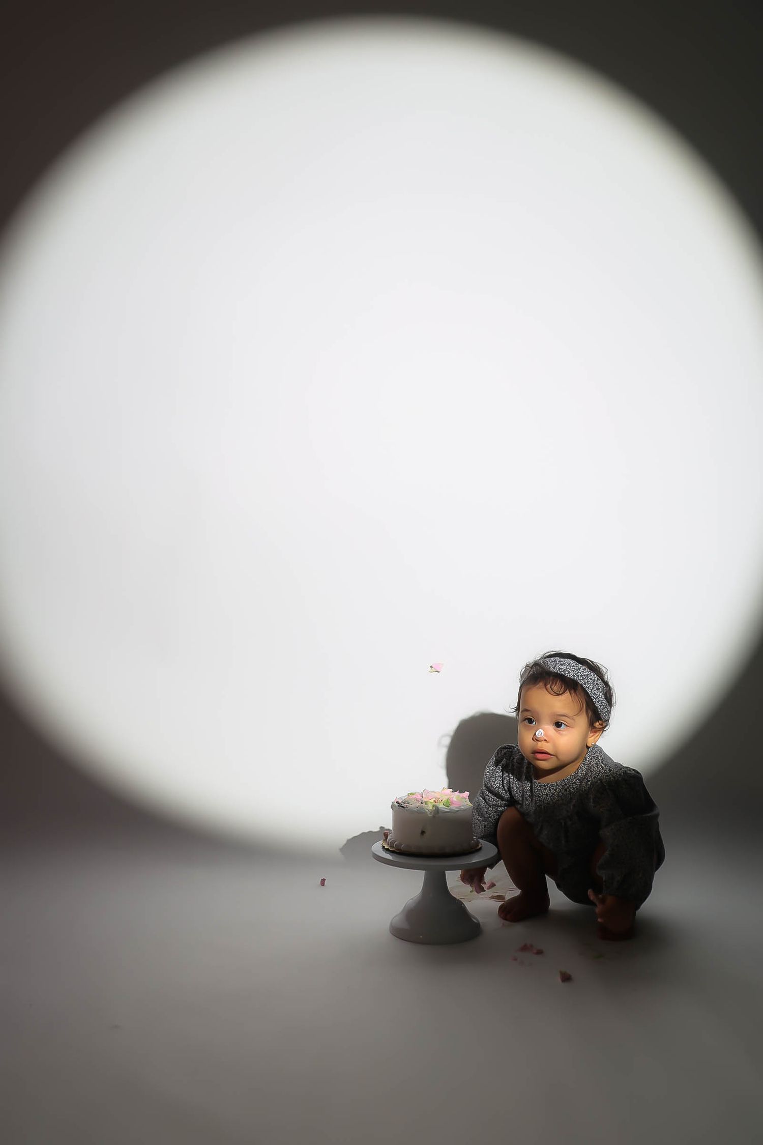 A baby in gray outfit sits beside a small birthday cake under a circular spotlight on a gray background.
