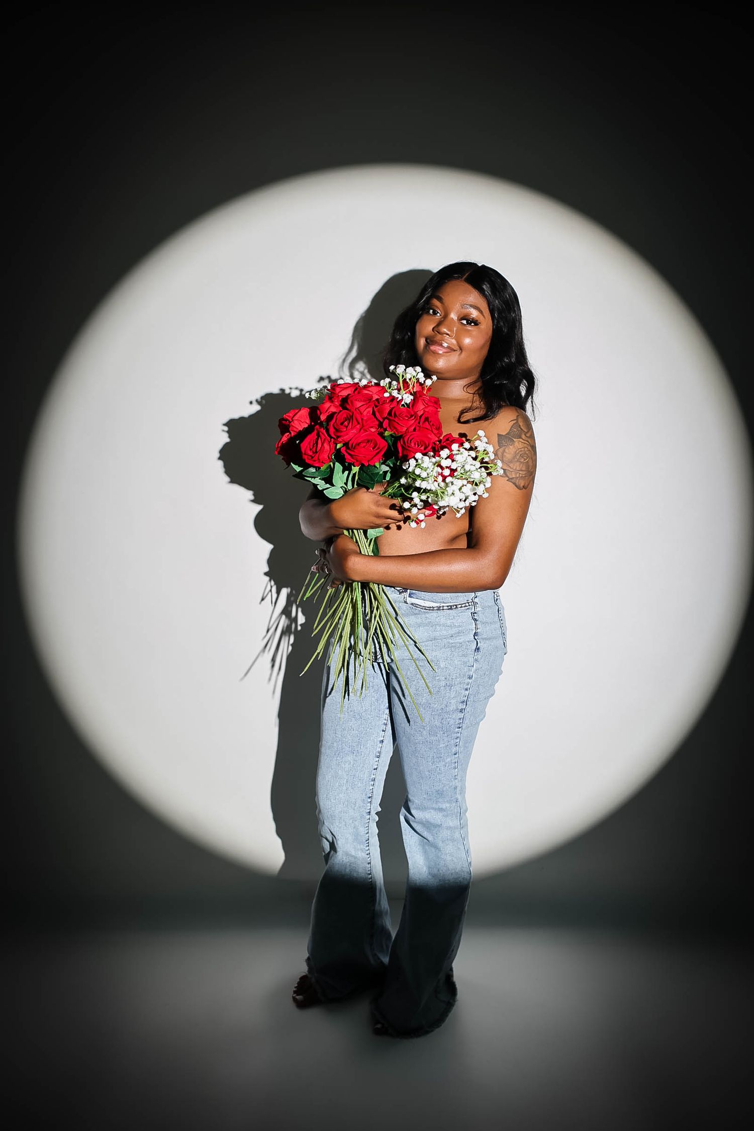 Smiling Black woman holding red roses bouquet in jeans and floral top, posed in spotlight on dark background.