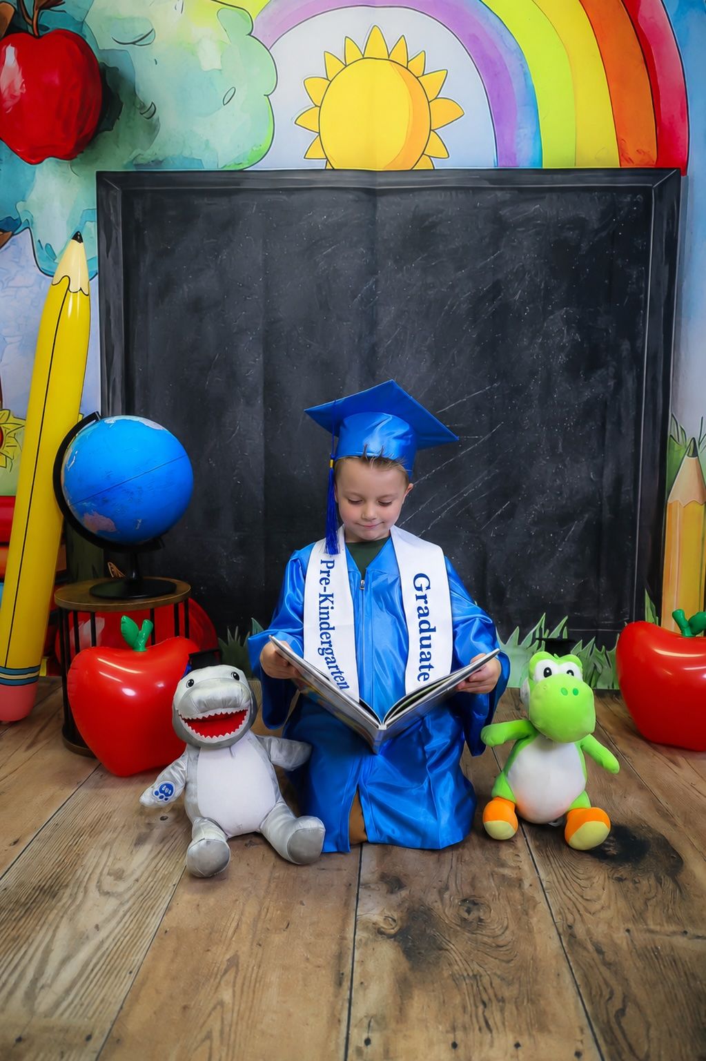 A young child in blue graduation cap and gown reads a book surrounded by stuffed animals and a chalkboard backdrop.