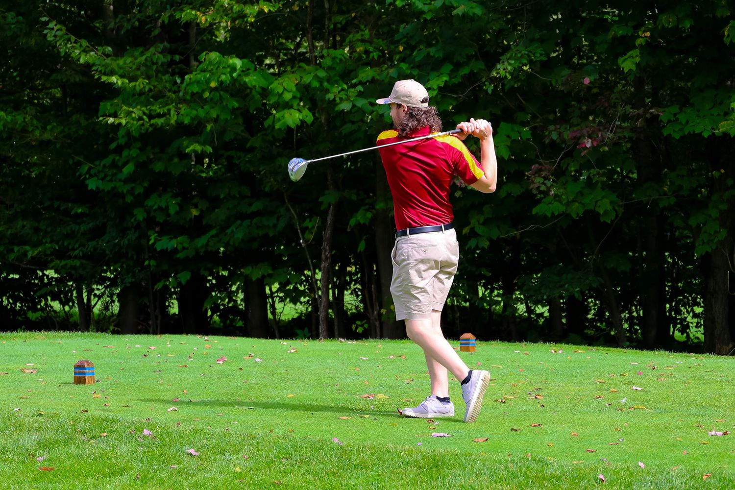 A golfer in a red shirt takes a swing on a lush green golf course surrounded by trees.