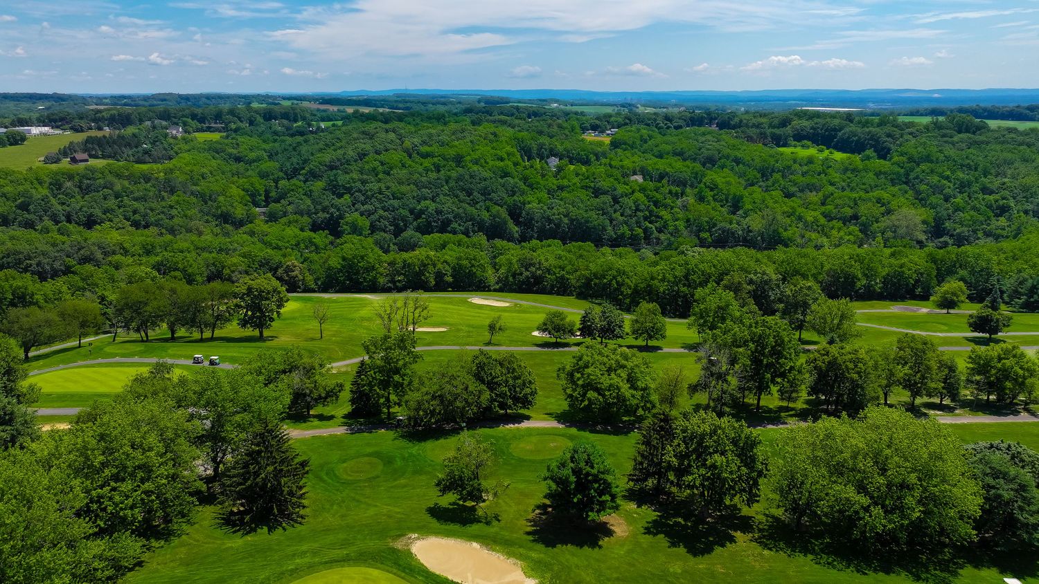 Aerial view of a lush green golf course nestled among rolling hills and forest on a sunny summer day.