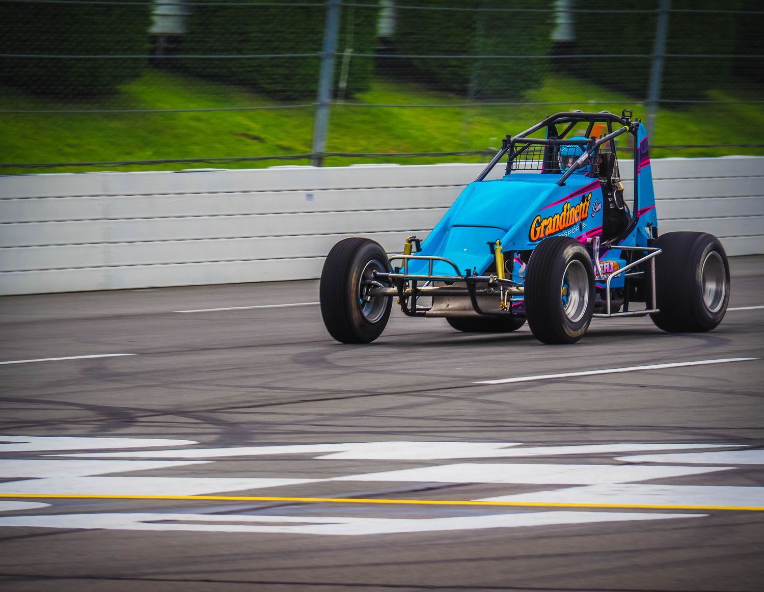 Race car speeds down wet racetrack during rainy conditions with spray trailing behind.