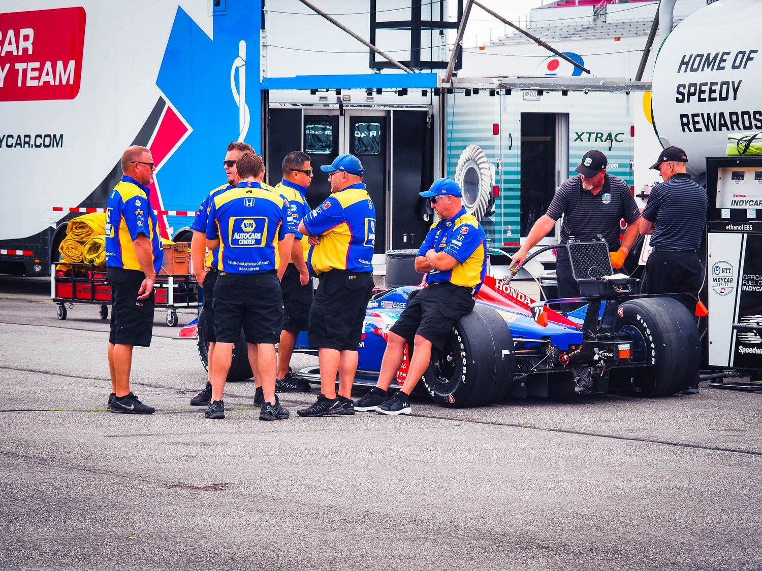 Racing pit crew members in blue uniforms gather around a race car during a motorsport event at the track.