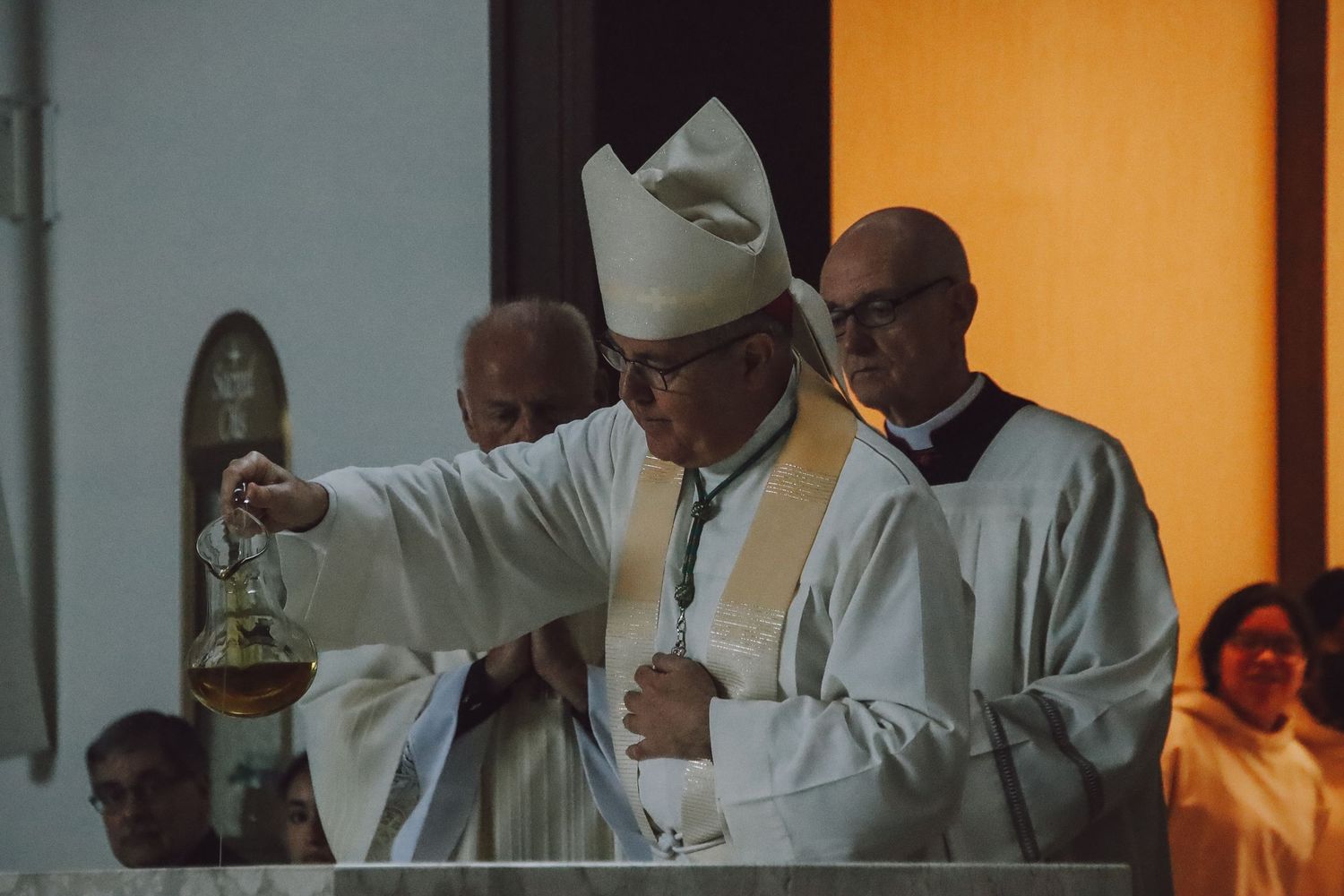 Priests in white vestments stand together during a church service with orange lighting in the background.