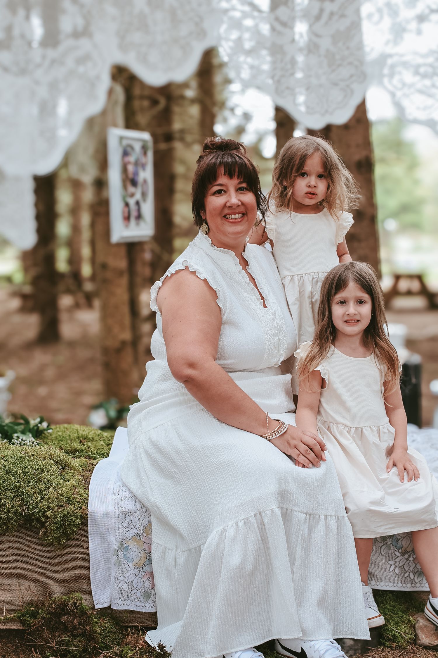 A group wearing coordinating white dresses poses together in a wooded outdoor setting for a family photo session.
