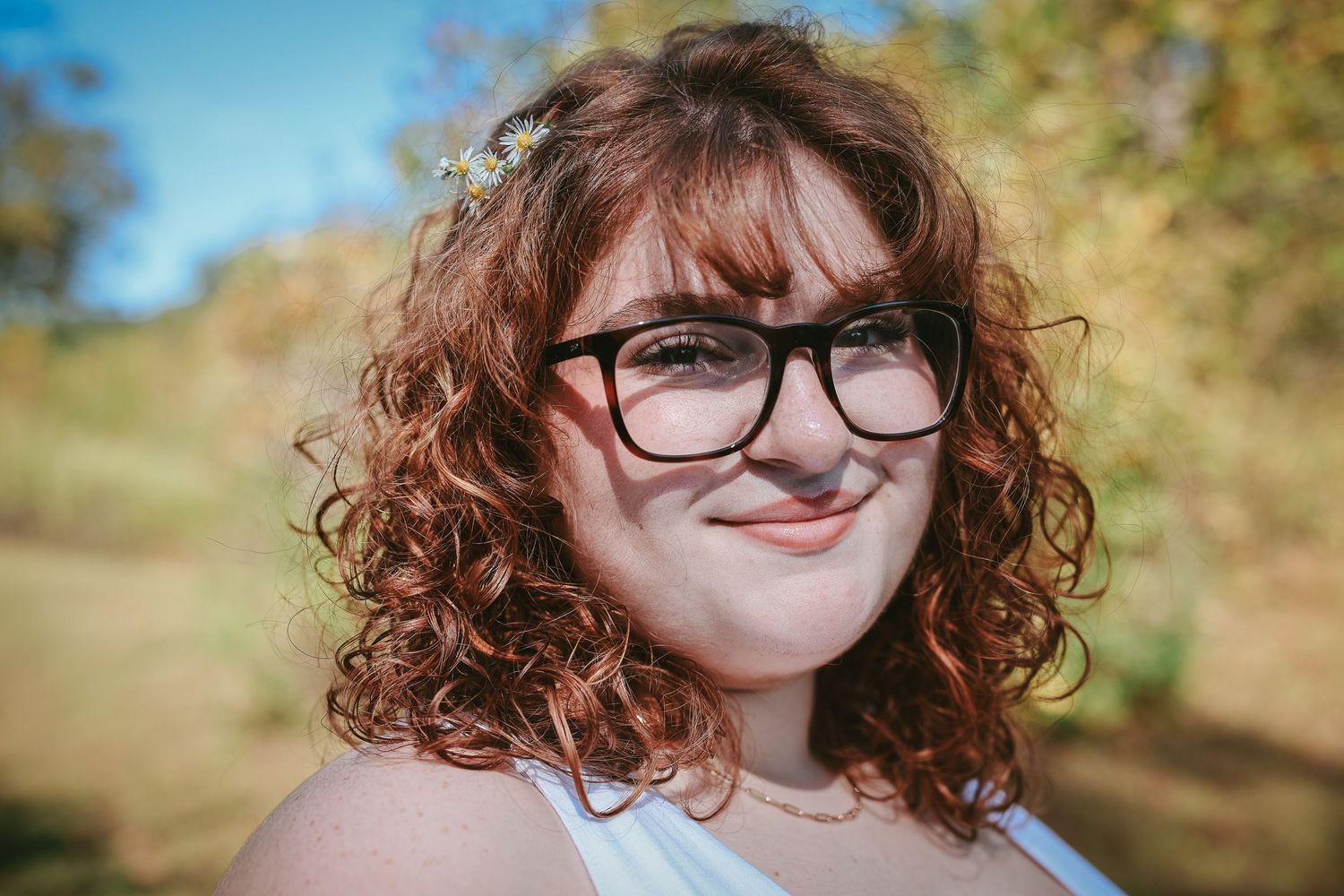 A close-up portrait of someone with curly brown hair and black-rimmed glasses smiling outdoors on a sunny day.